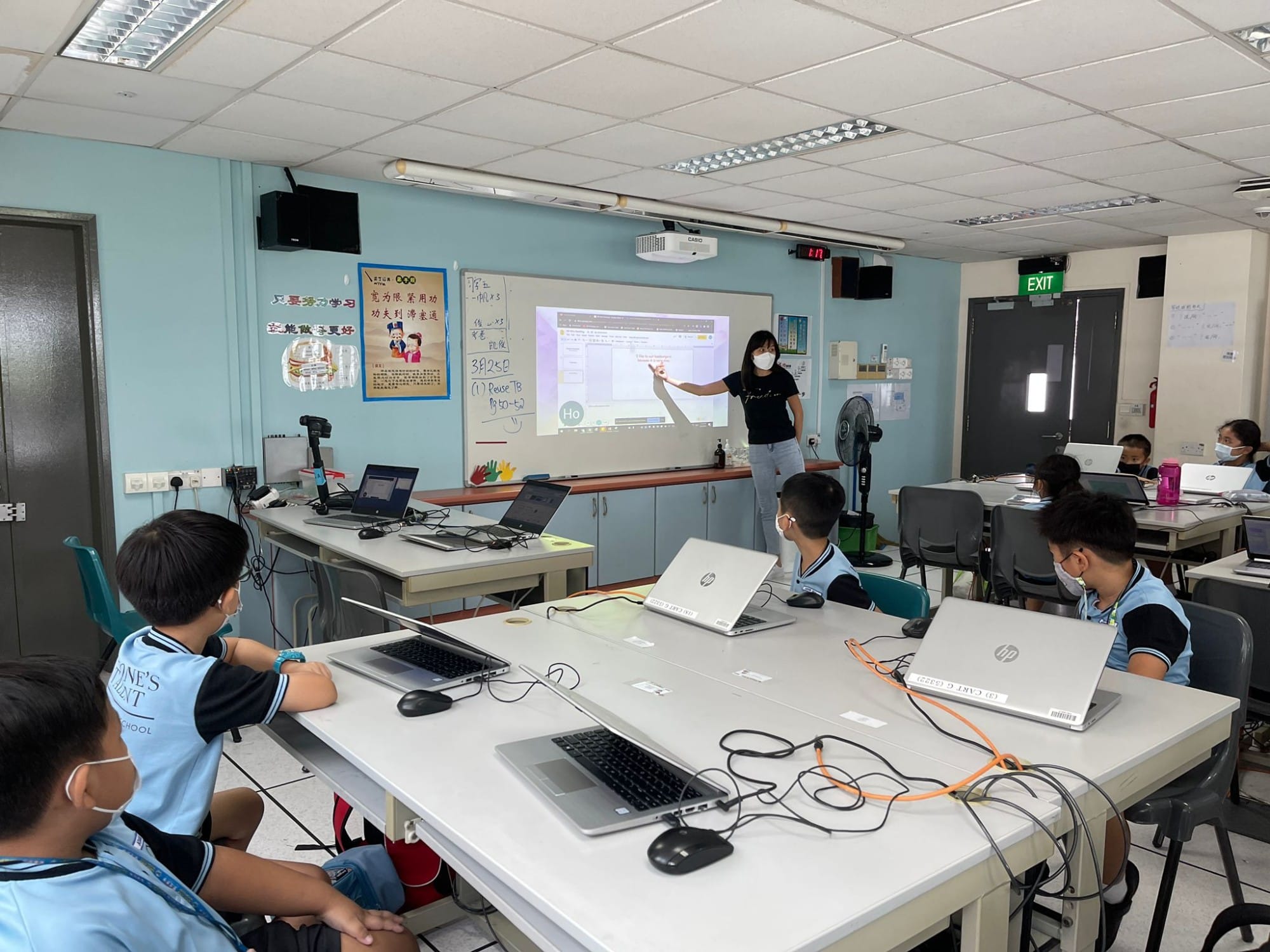 A masked teacher points at a projected screen while masked students sit at tables with laptops in a classroom.