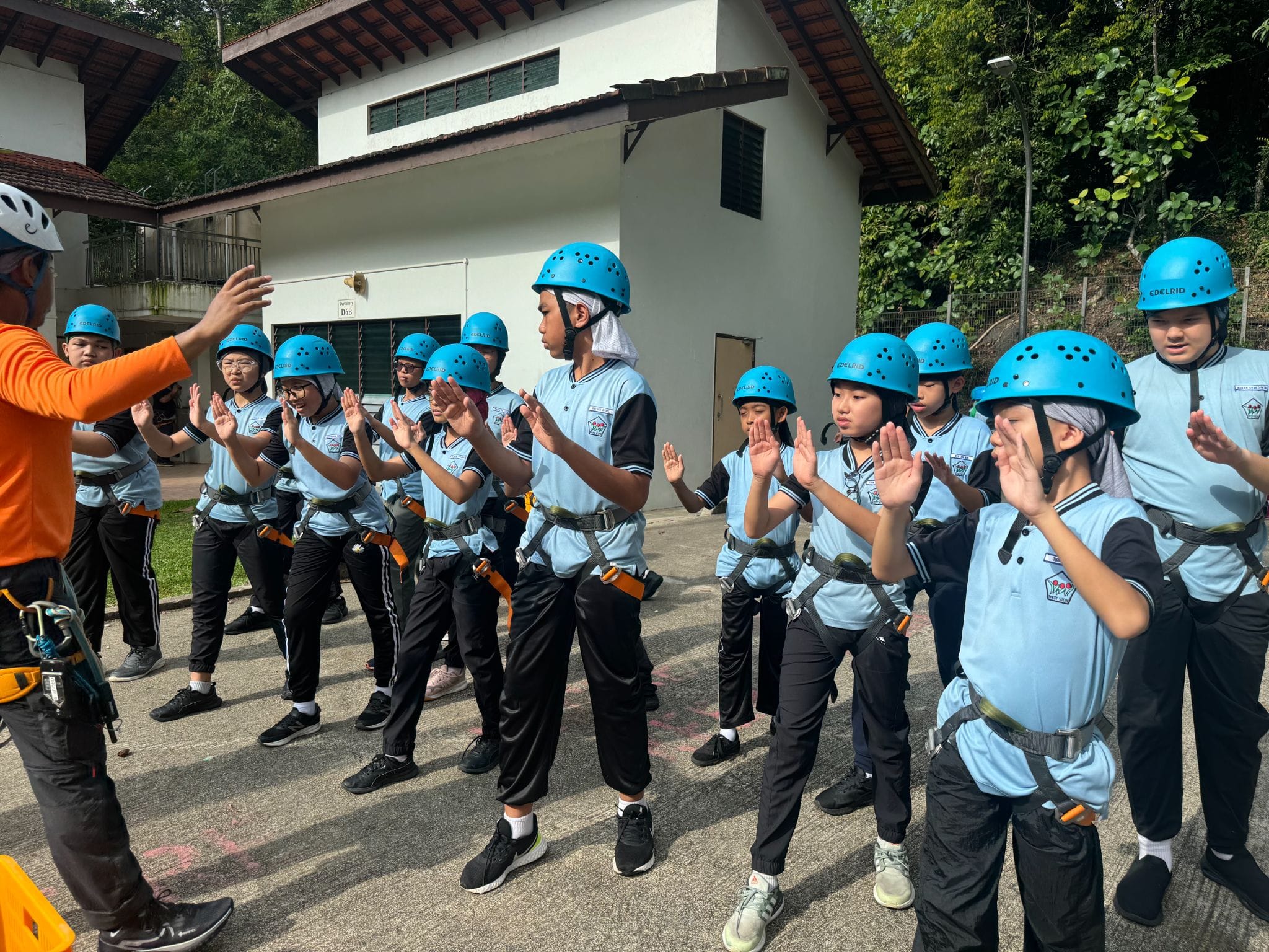 Students in blue helmets and harnesses raise their hands outdoors while an instructor in orange guides them.
