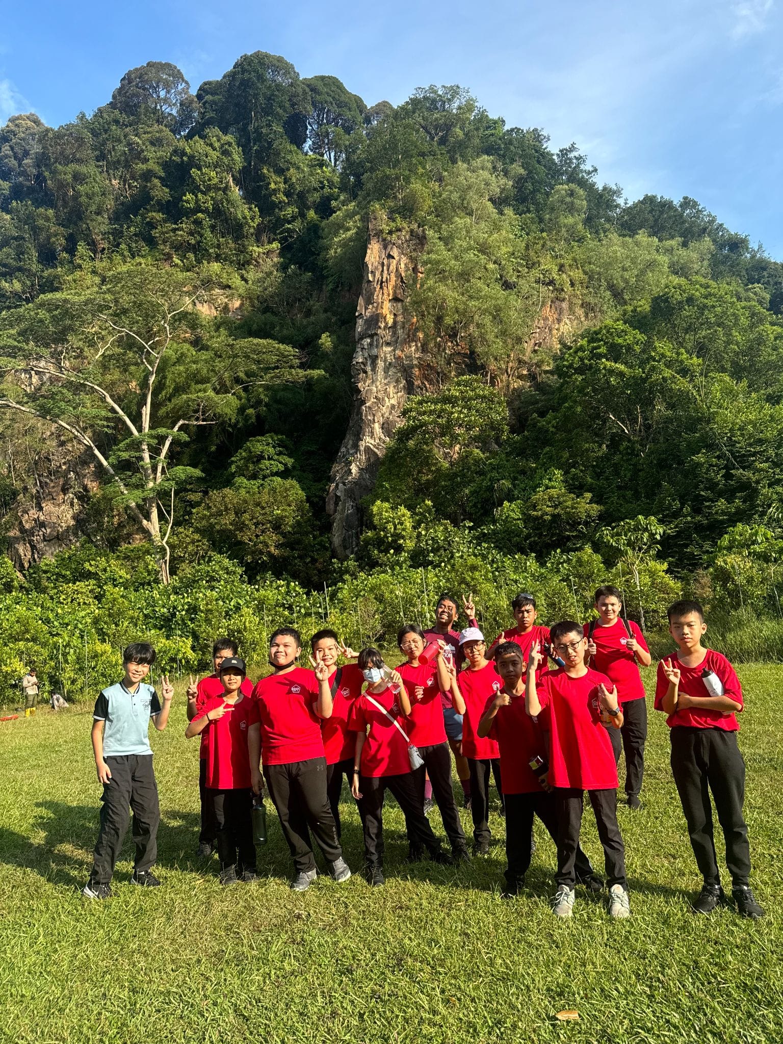 A group of young people mostly in red shirts pose on grass before a tall rocky cliff covered with dense green trees.