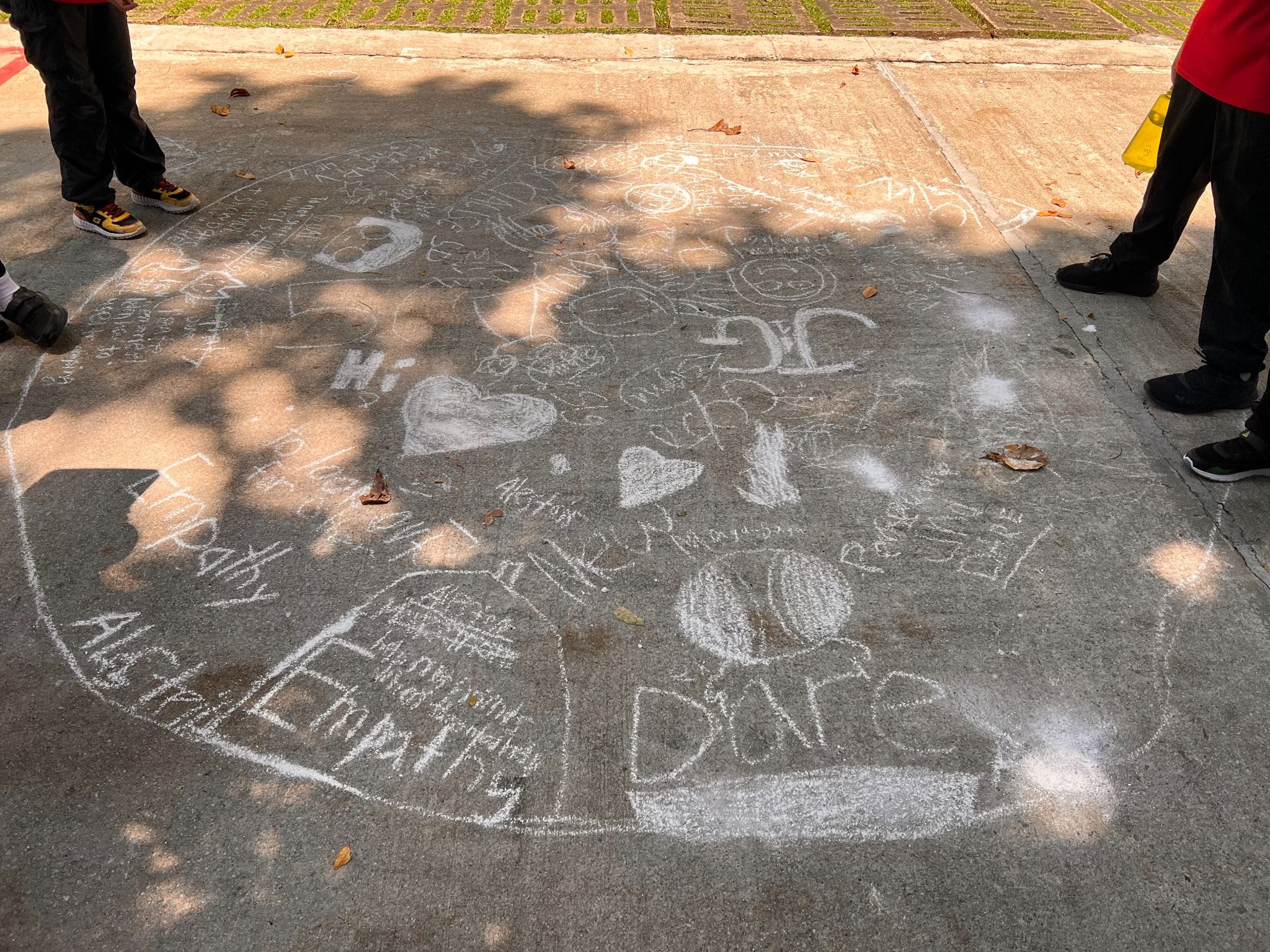 Sidewalk covered in white chalk drawings and words like Empathy, Dare, and Hi, with children's legs visible around it.