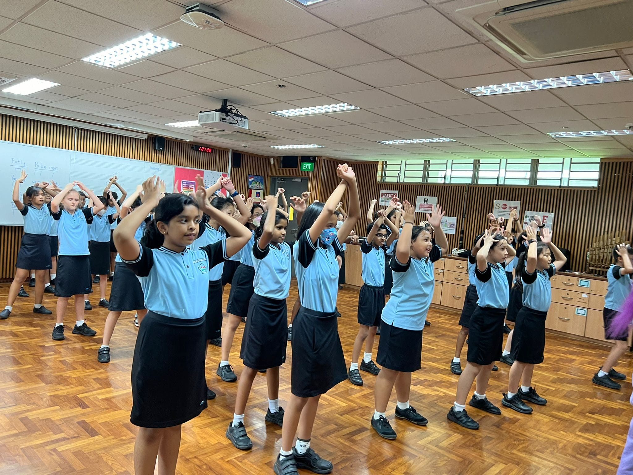 Girls in blue and black school uniforms raise their arms during a group activity in a wood-paneled hall.