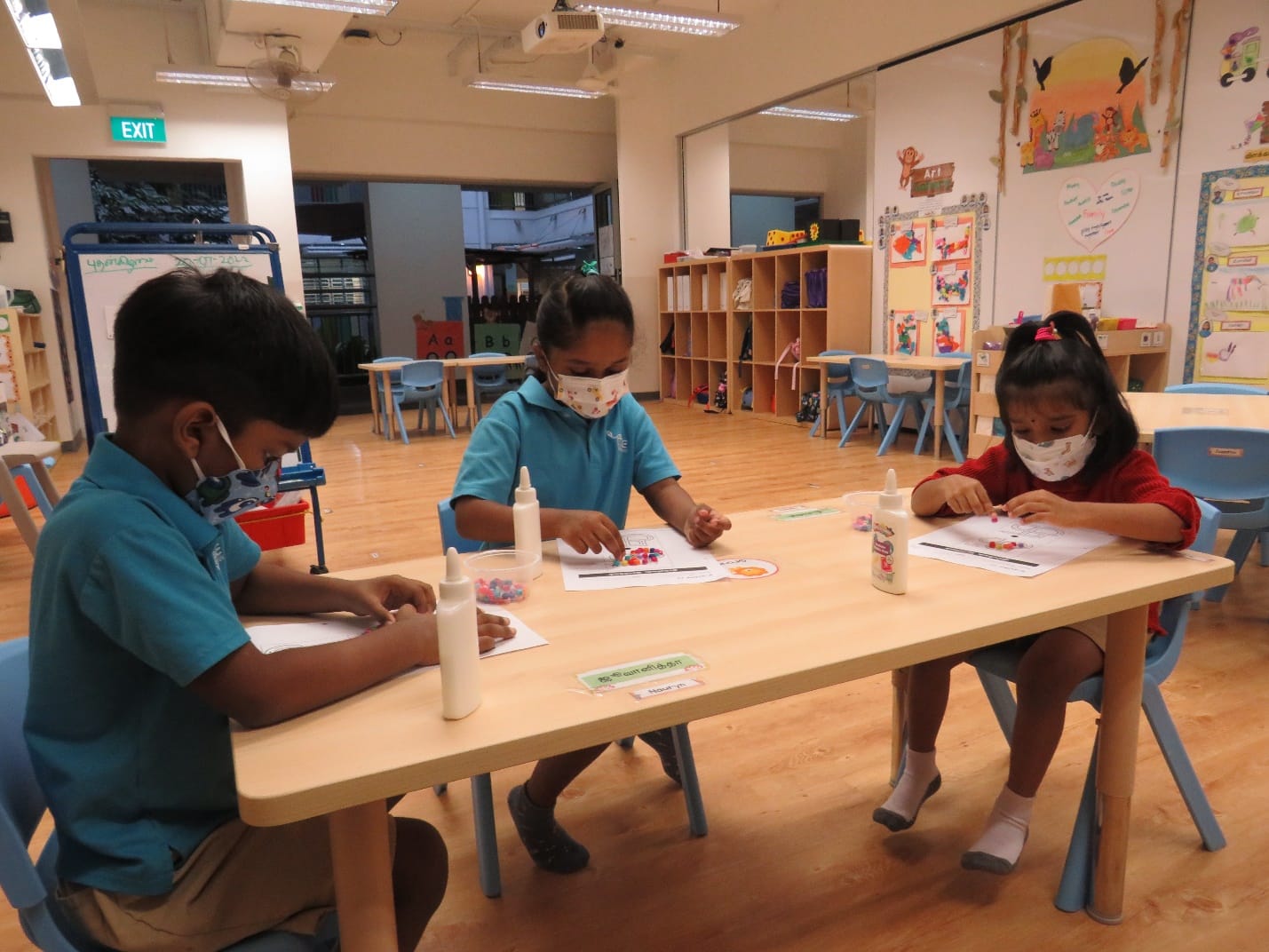 Three young children wearing face masks do a craft activity with glue at a table in a colorful classroom.