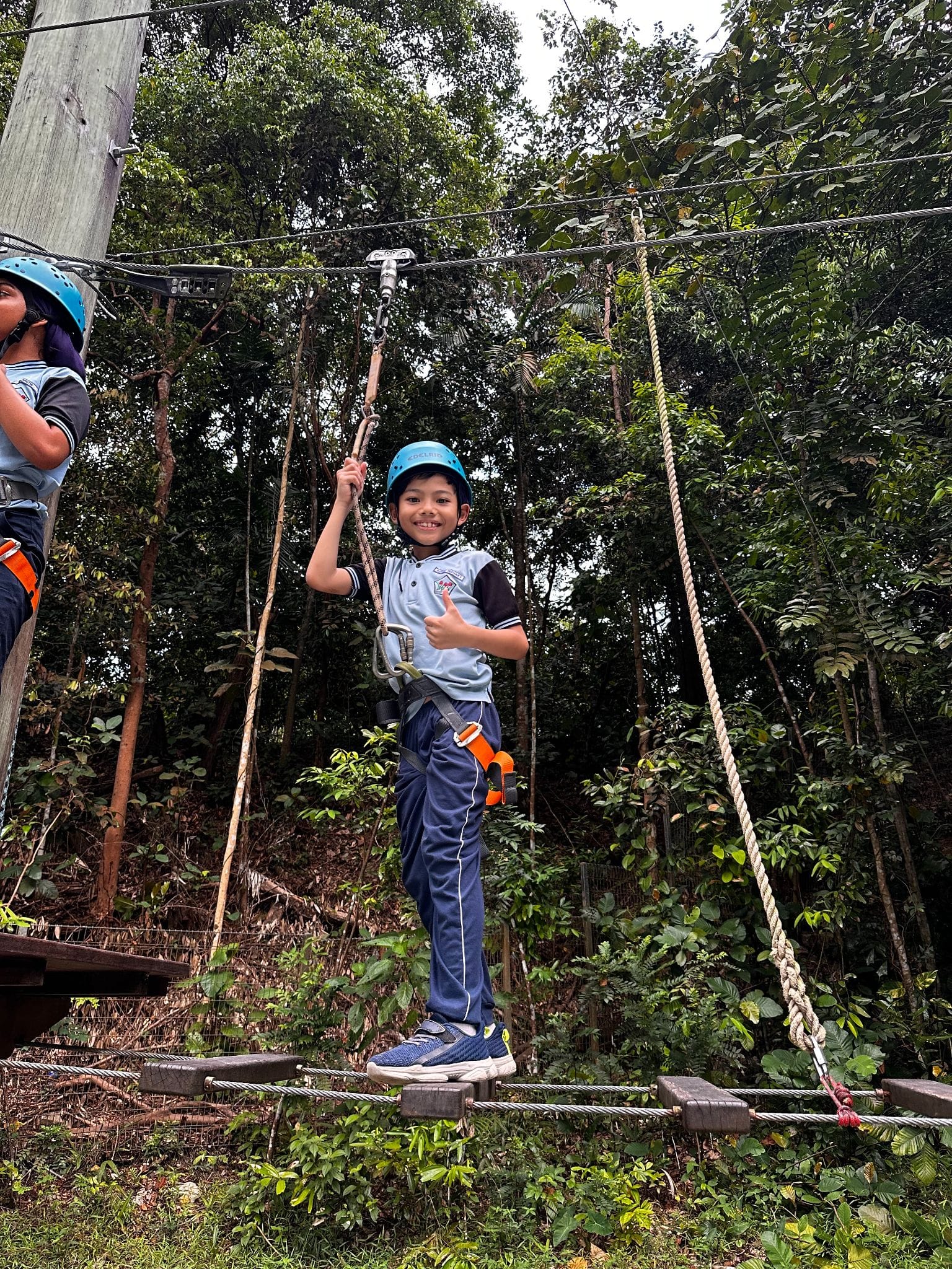 A smiling boy wearing a blue helmet and harness gives a thumbs up while standing on a rope bridge in a forest.