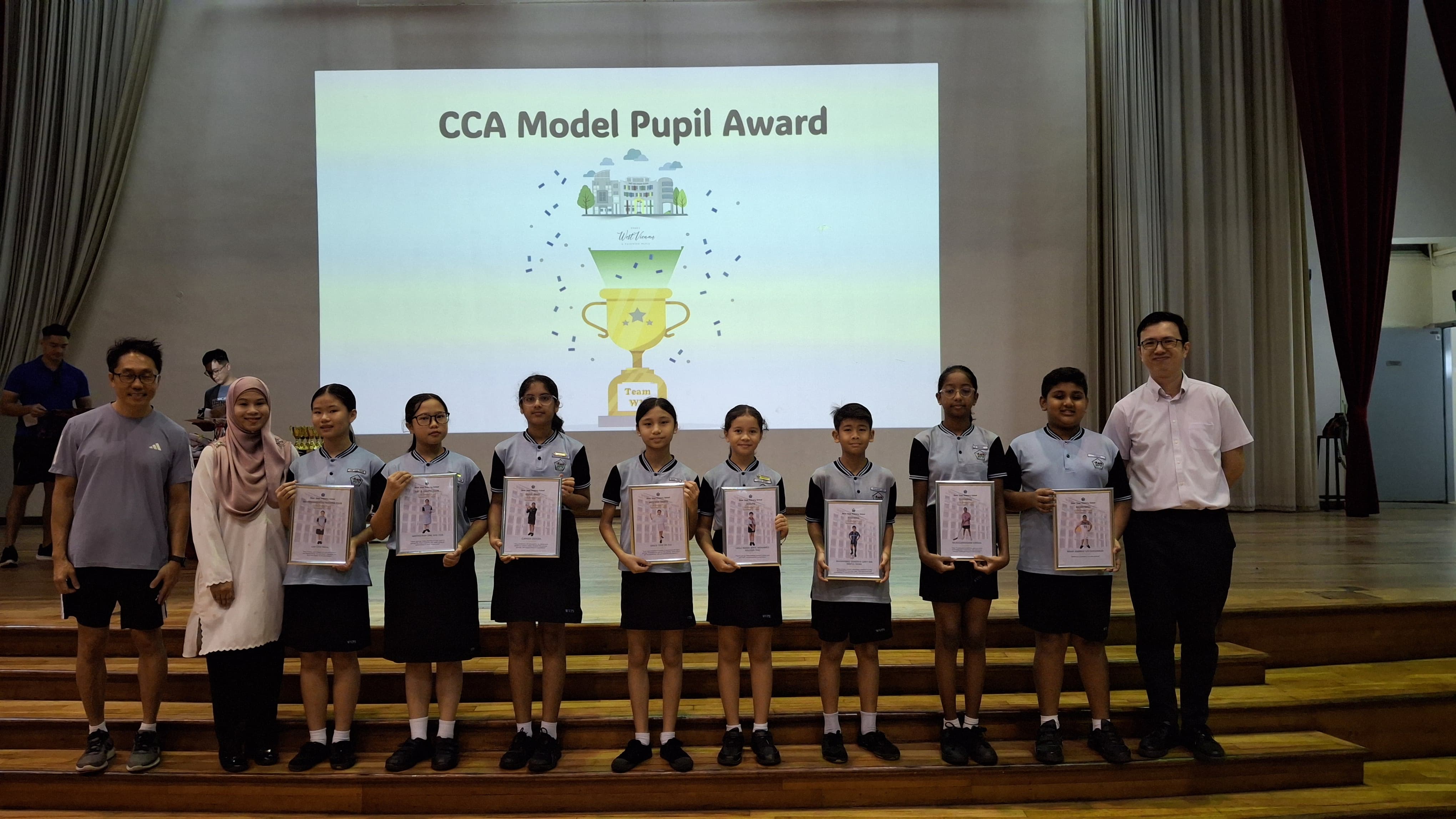 Students in school uniforms hold framed certificates on a stage under a CCA Model Pupil Award projection screen.