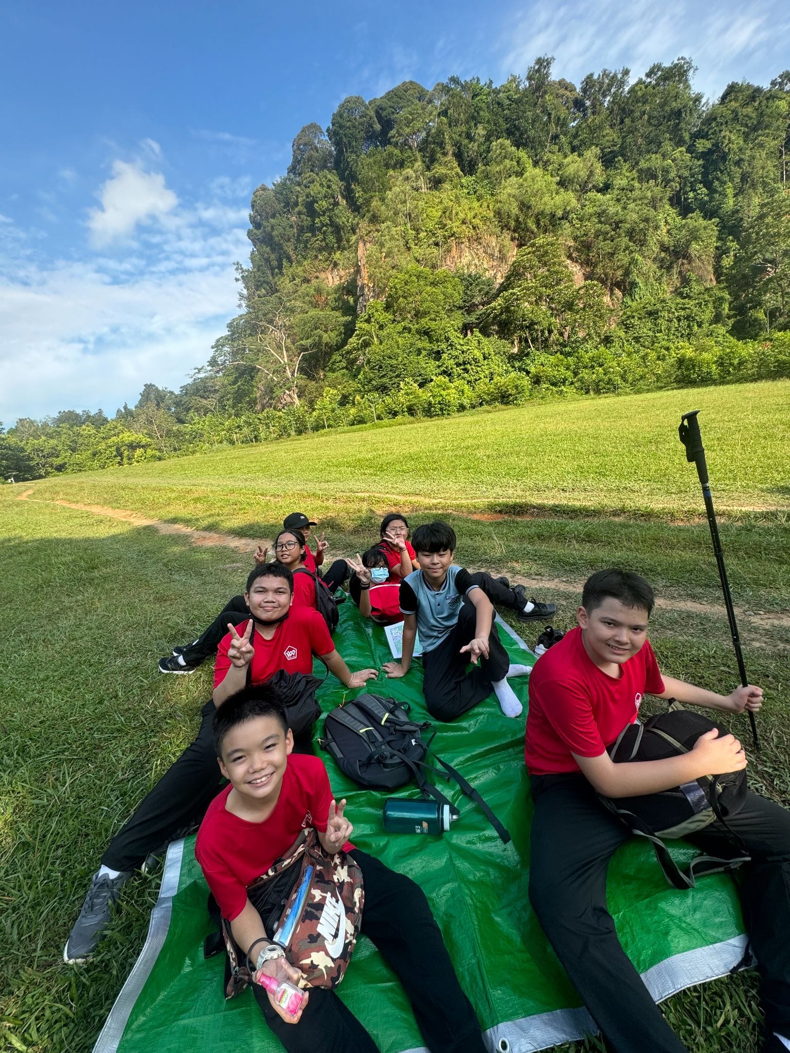 Six children in red shirts sit and lie on a green tarp on grass, posing with peace signs before a forested rocky hill.