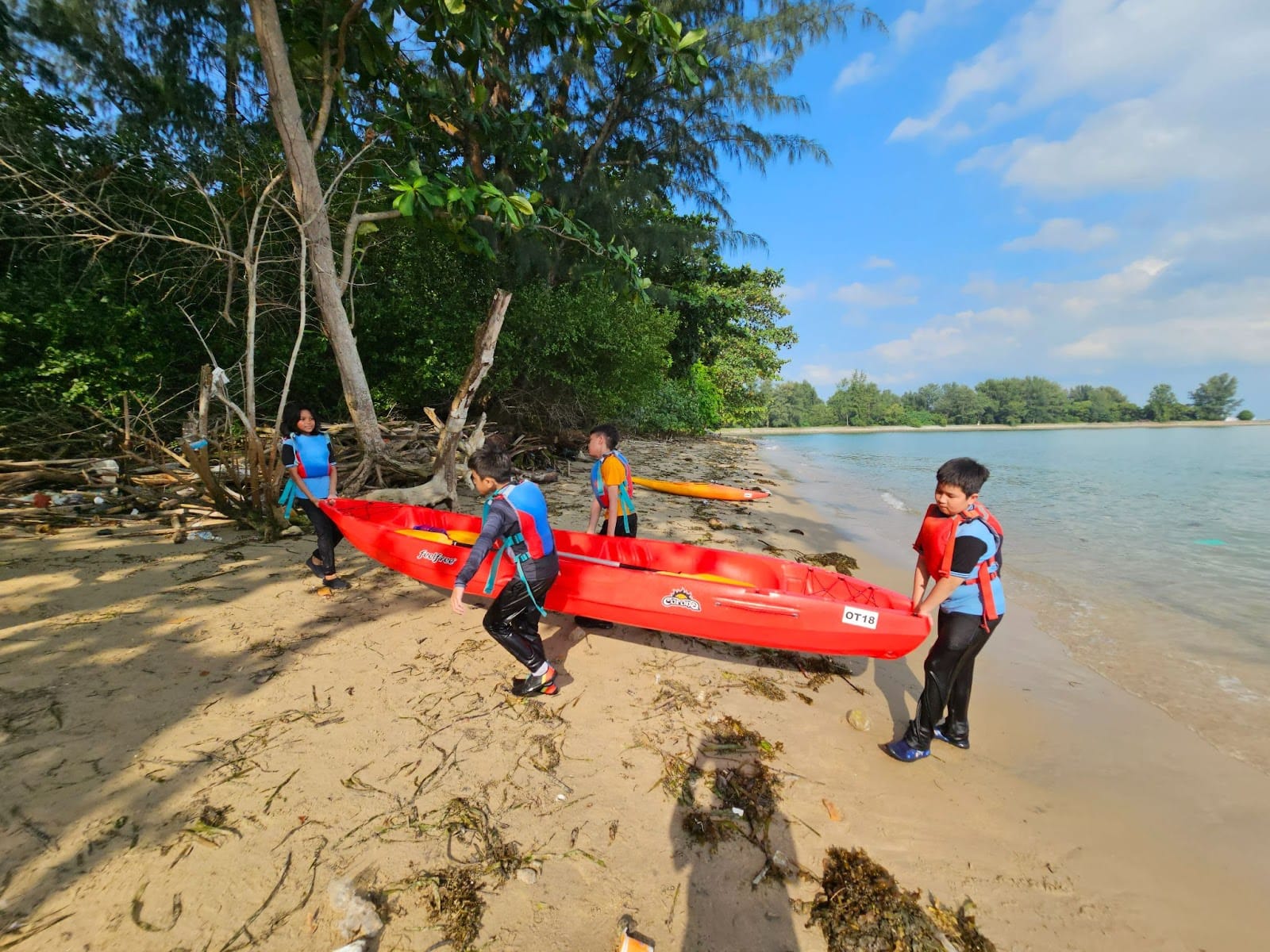 Four children in life vests carry a red kayak along a sandy beach toward the water.