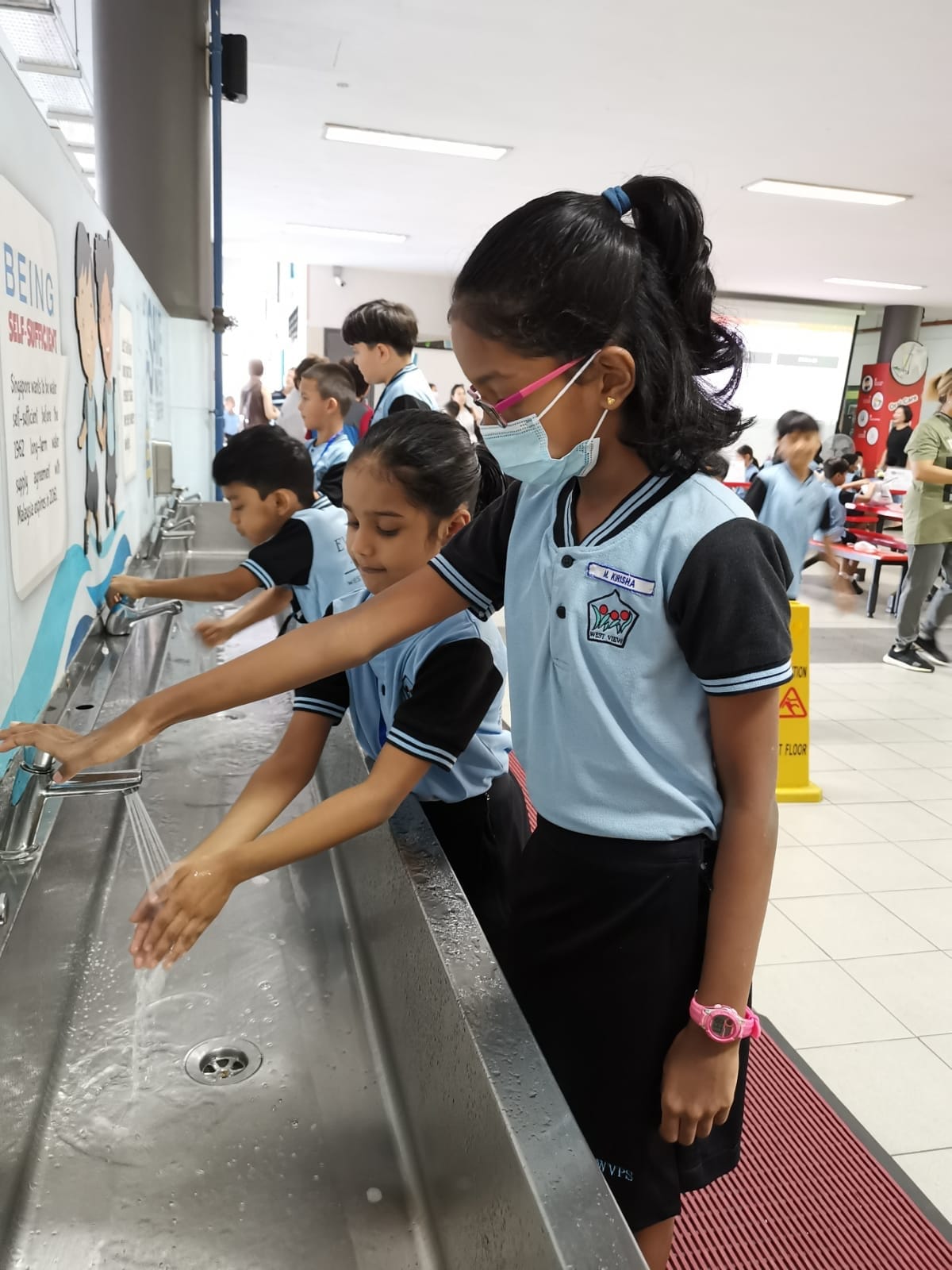 School children in blue uniforms washing hands at a row of metal sinks, one girl wearing a face mask.
