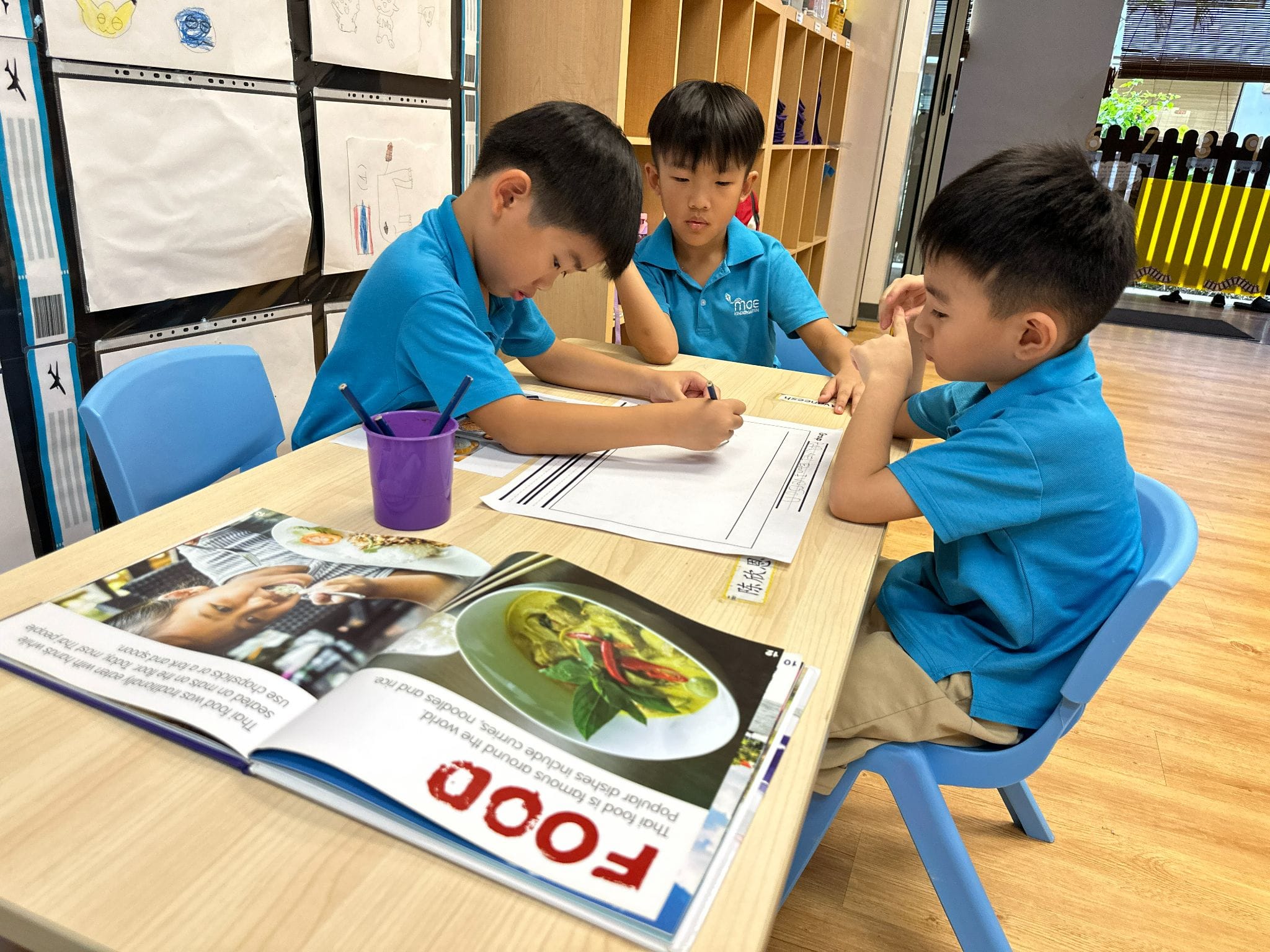 Three young boys in blue uniforms sit at a classroom table with an open food book and a worksheet.