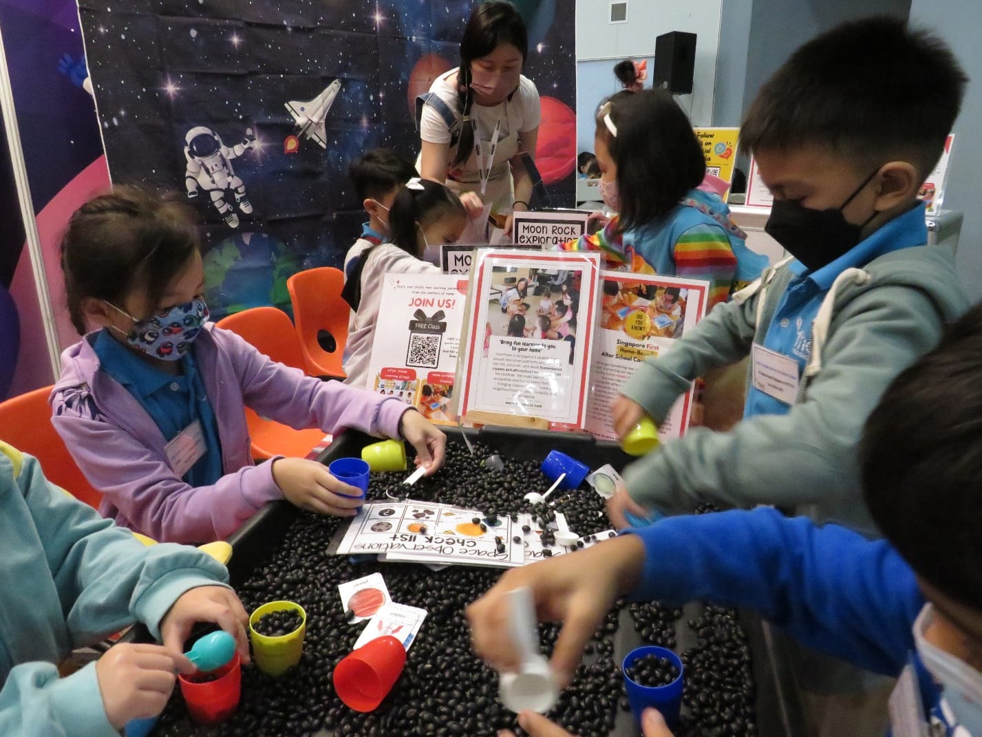 Children in masks gather around a table covered in dark beads for a Moon Rock Exploration activity with colorful cups.