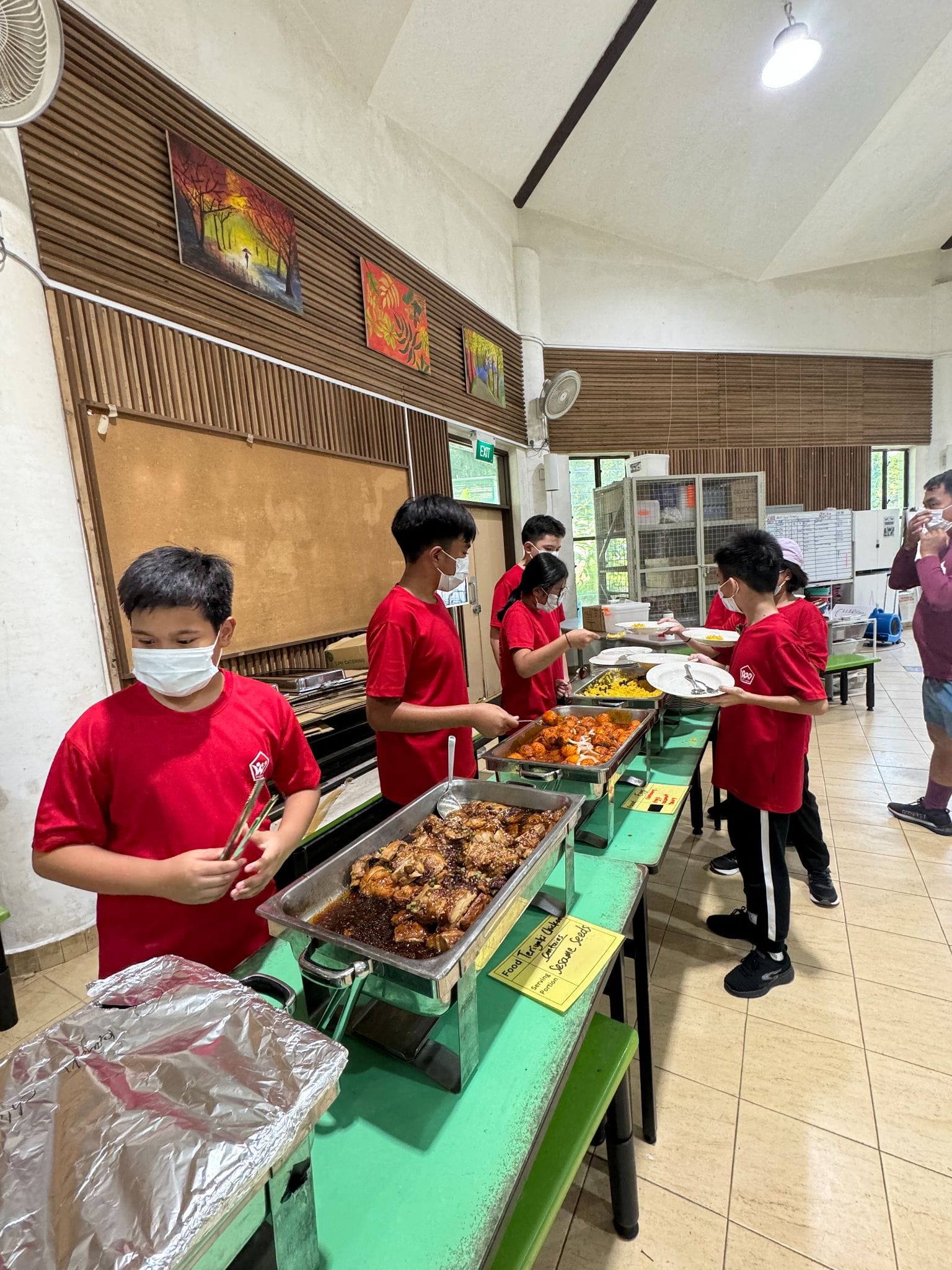 Children in red shirts and face masks serve food from buffet trays in a school hall.