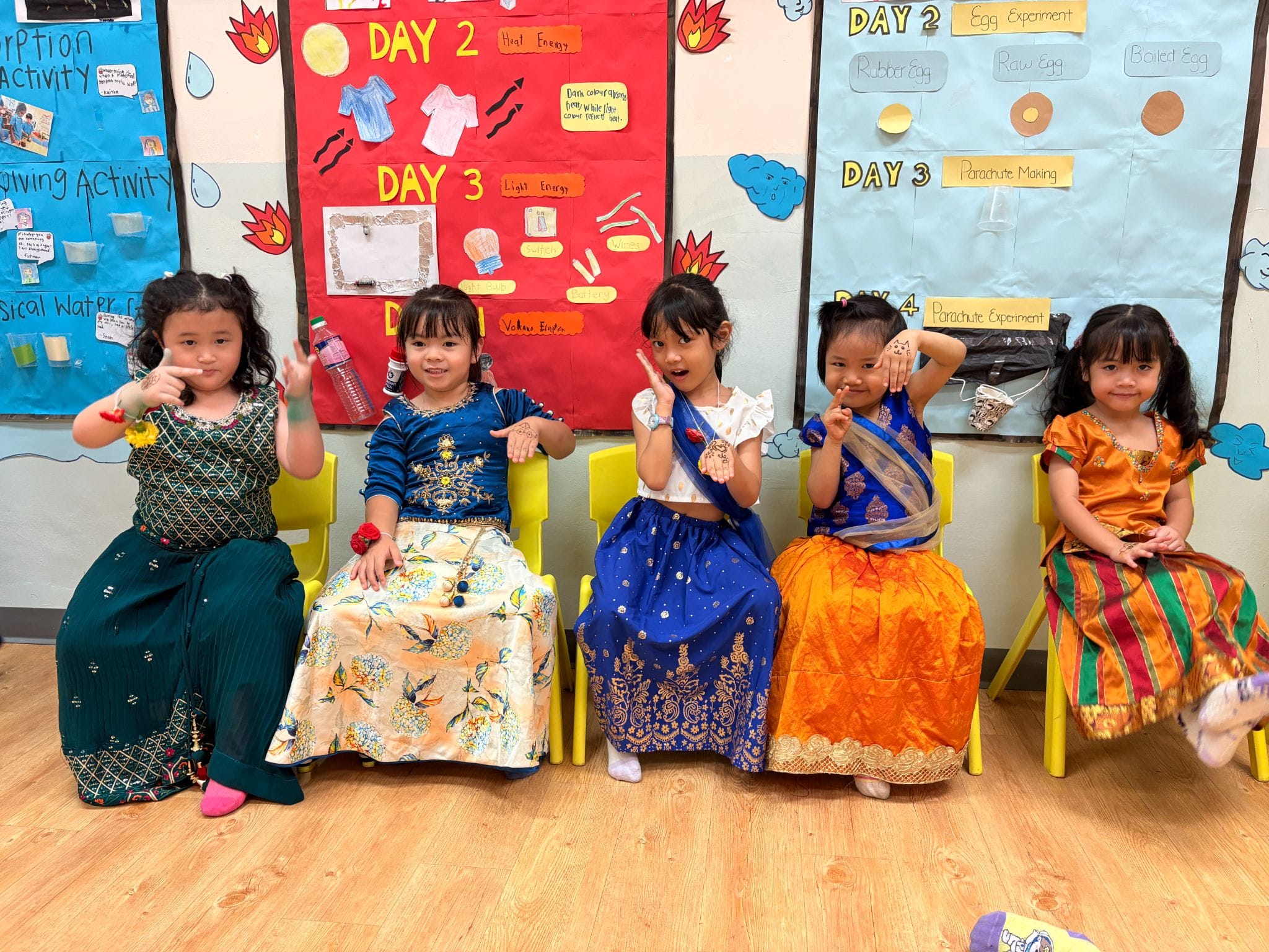 Five young girls in colorful traditional outfits sit on yellow chairs in a classroom with science activity posters.