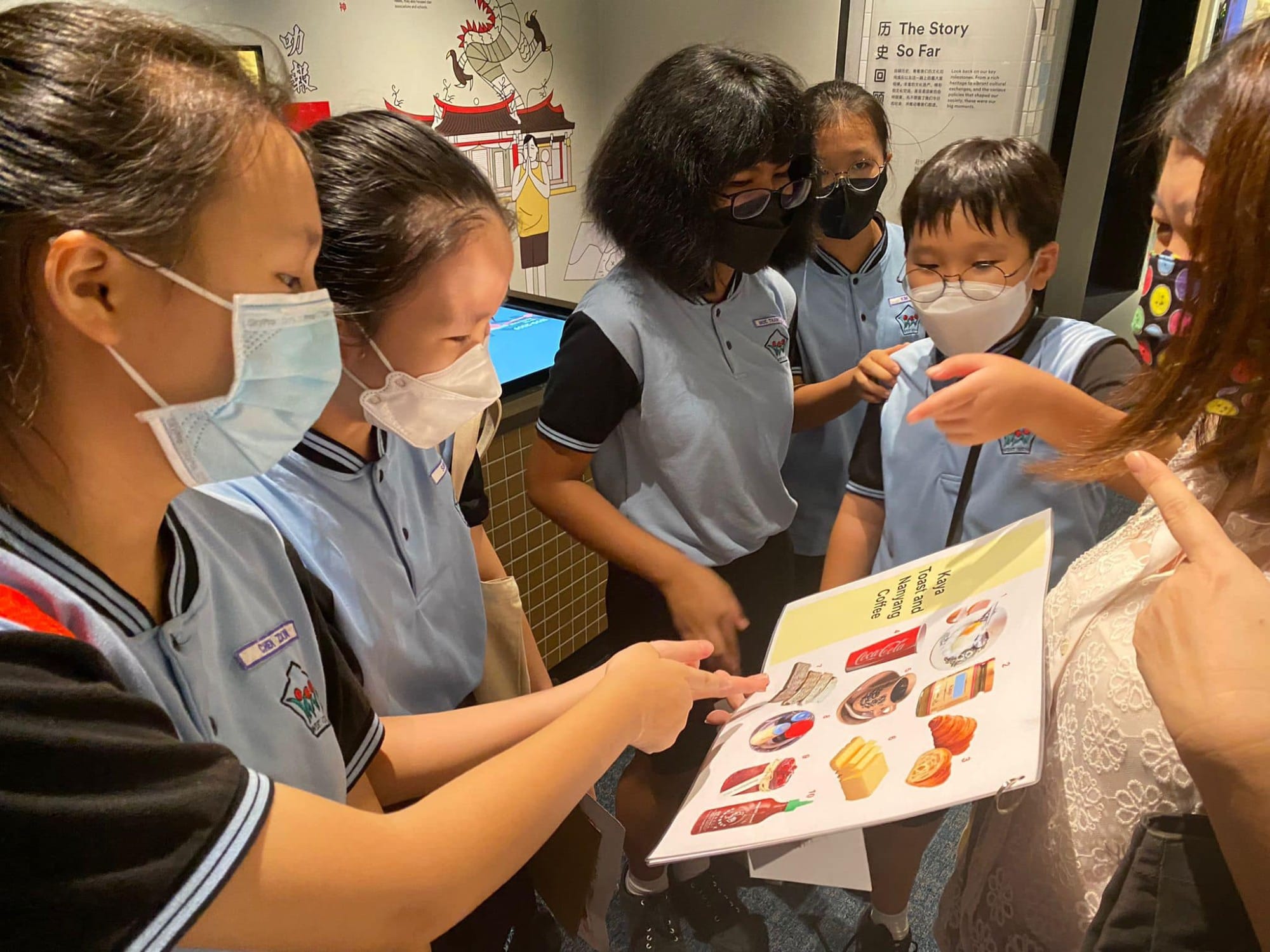 Five masked school children in blue uniforms look at a colorful booklet held by an adult in a museum.