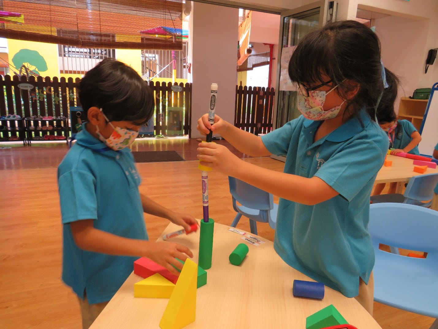 Two young masked children in teal uniforms stack colorful foam blocks and markers on a classroom table.