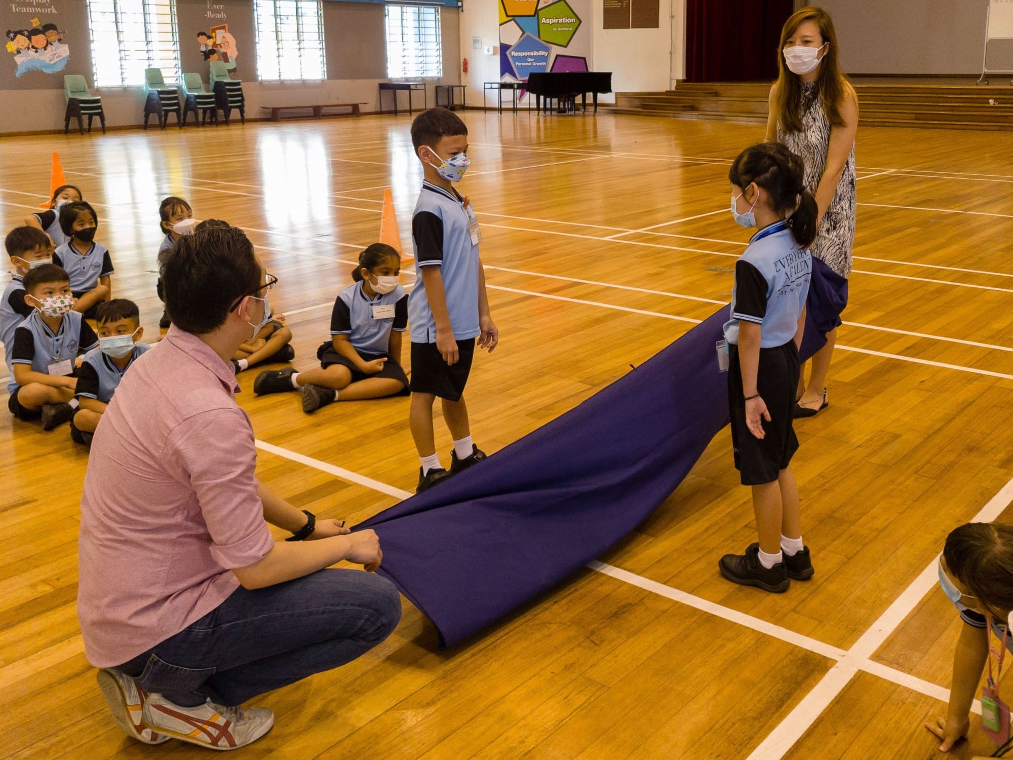 Masked children in school uniforms stand on a gym floor as an adult holds a purple cloth during an activity.