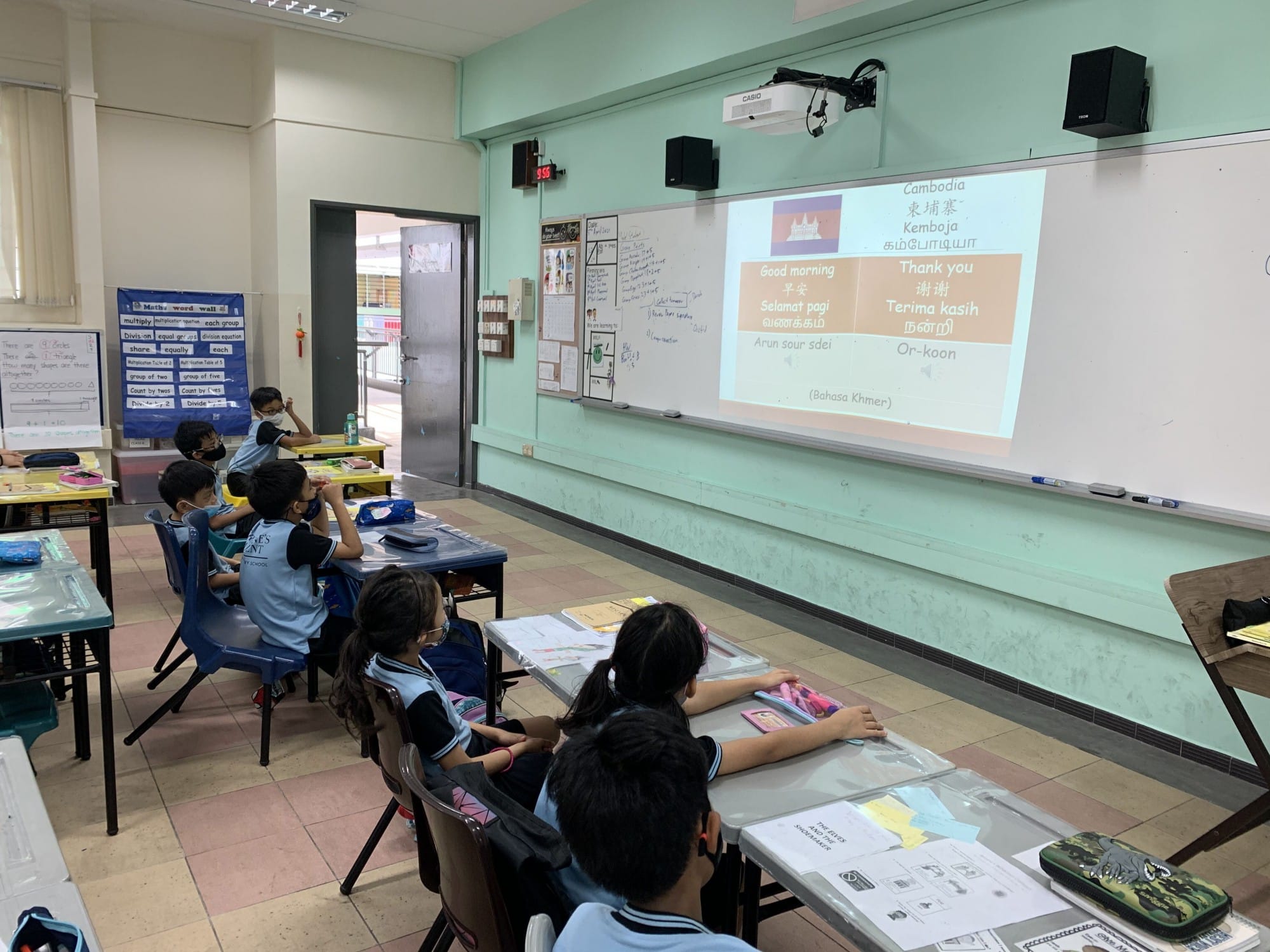 Young students in a classroom watch a projected slide about Cambodia showing greetings in multiple languages.