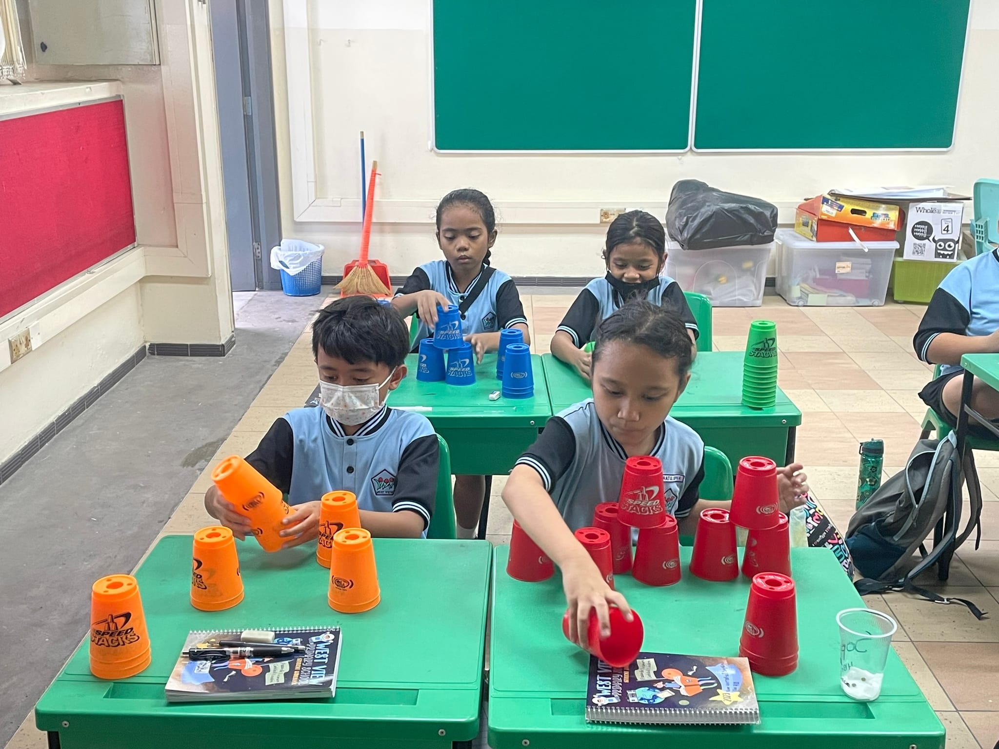 Four children in school uniforms stack colorful Speed Stacks cups on green desks in a classroom.