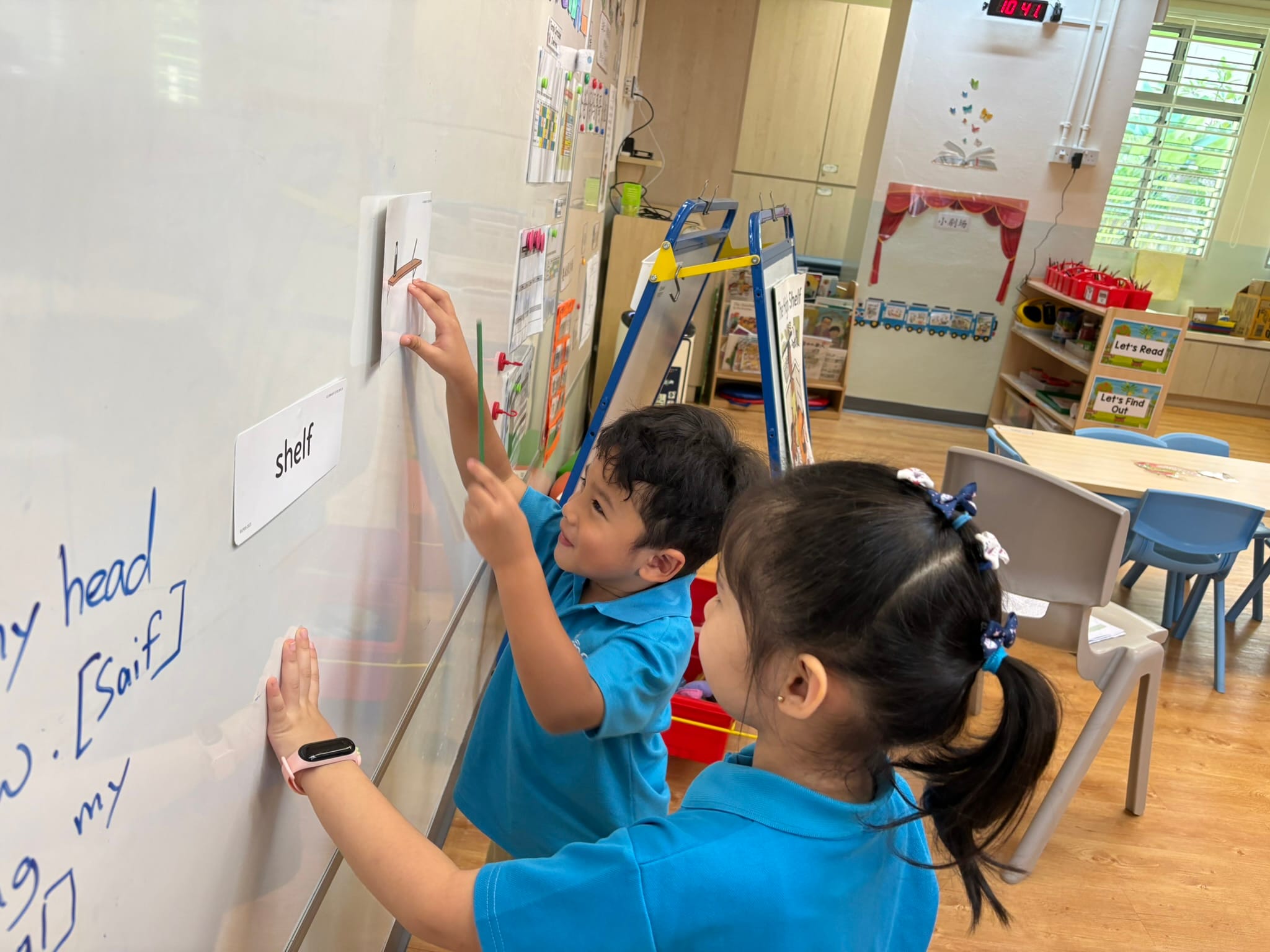 Two young children in blue uniforms point at word cards on a whiteboard in a colorful classroom.