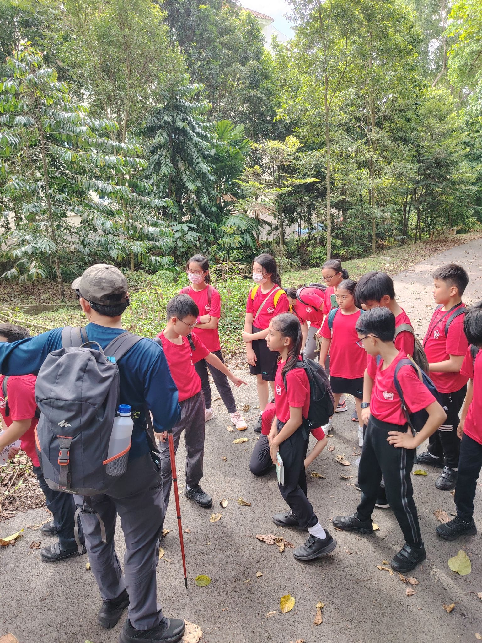 A group of children in red shirts with backpacks gather on a path surrounded by green trees, listening to an adult guide.