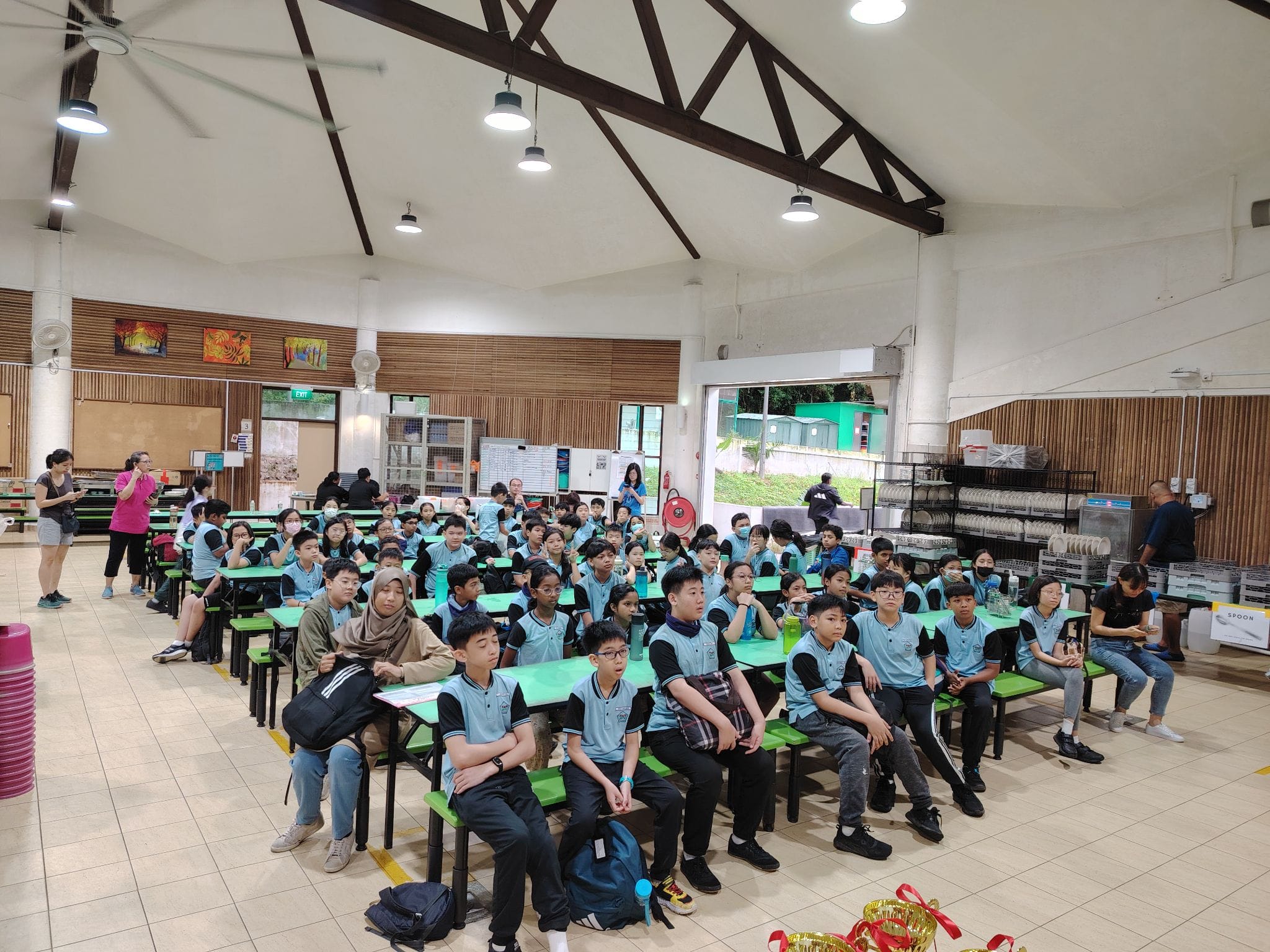 Students in blue uniforms sit at green tables in a school canteen, facing forward during an assembly.