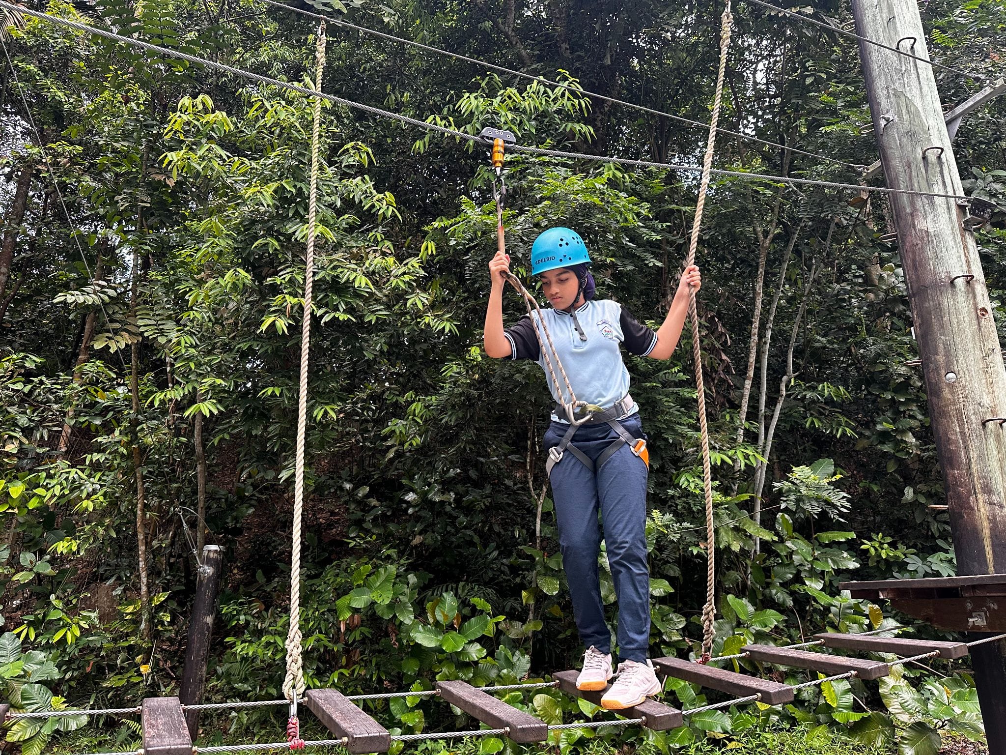 A young person wearing a blue helmet and harness navigates a rope and wooden plank obstacle course outdoors.