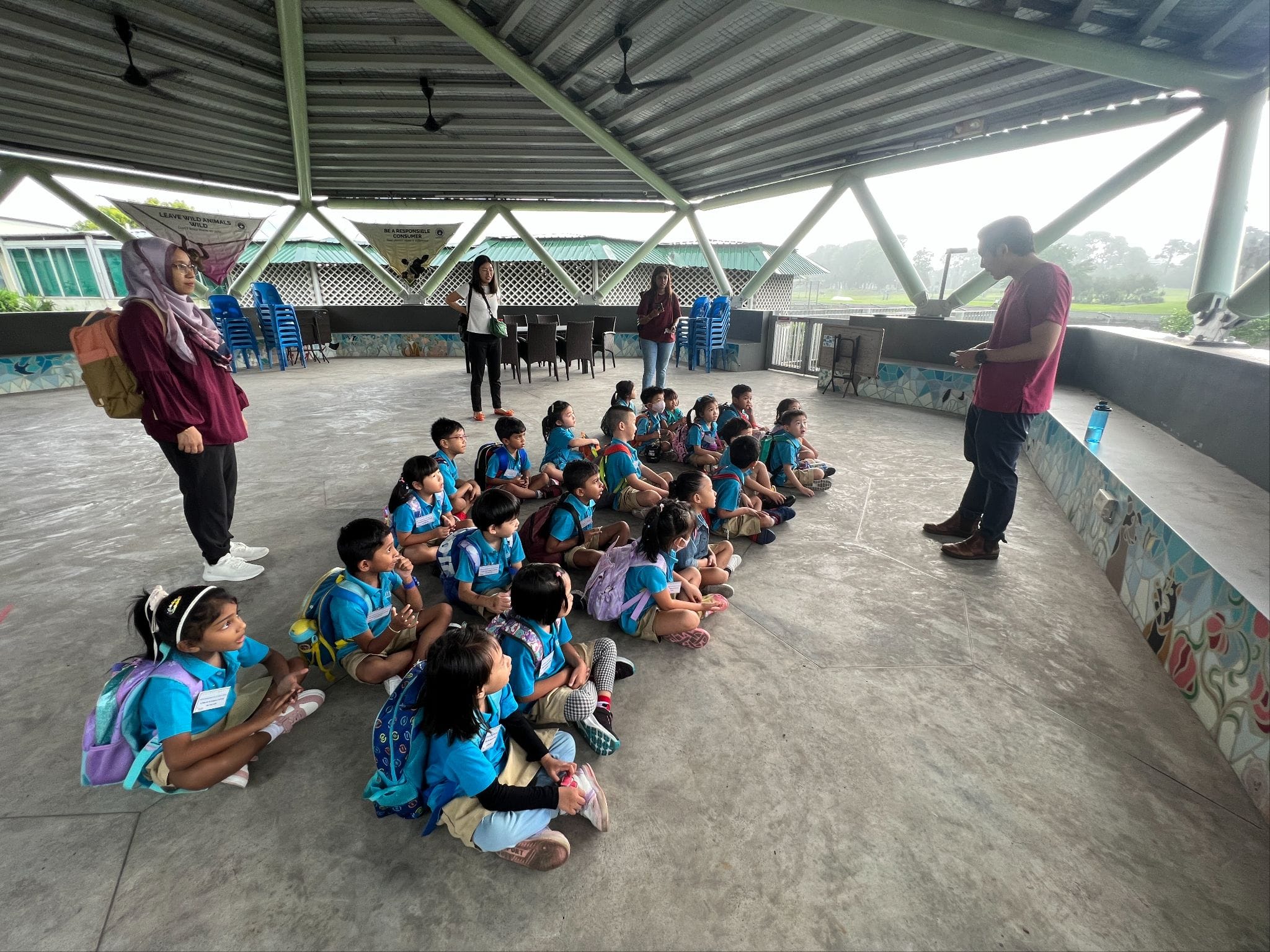A group of young students in blue uniforms sit on the floor under an open pavilion while adults stand around them.