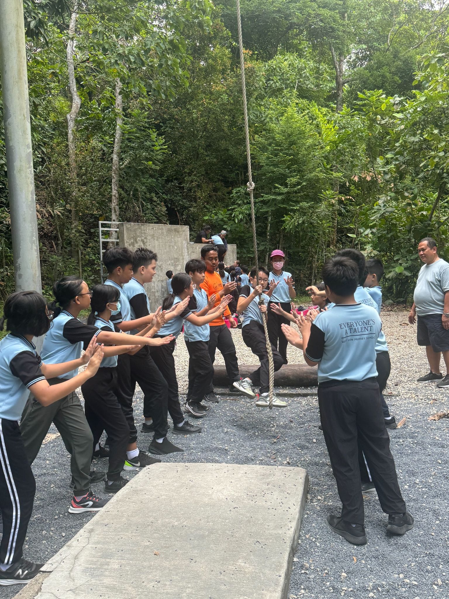 Students in light blue uniforms clap and cheer outdoors near a hanging rope and trees during a group activity.
