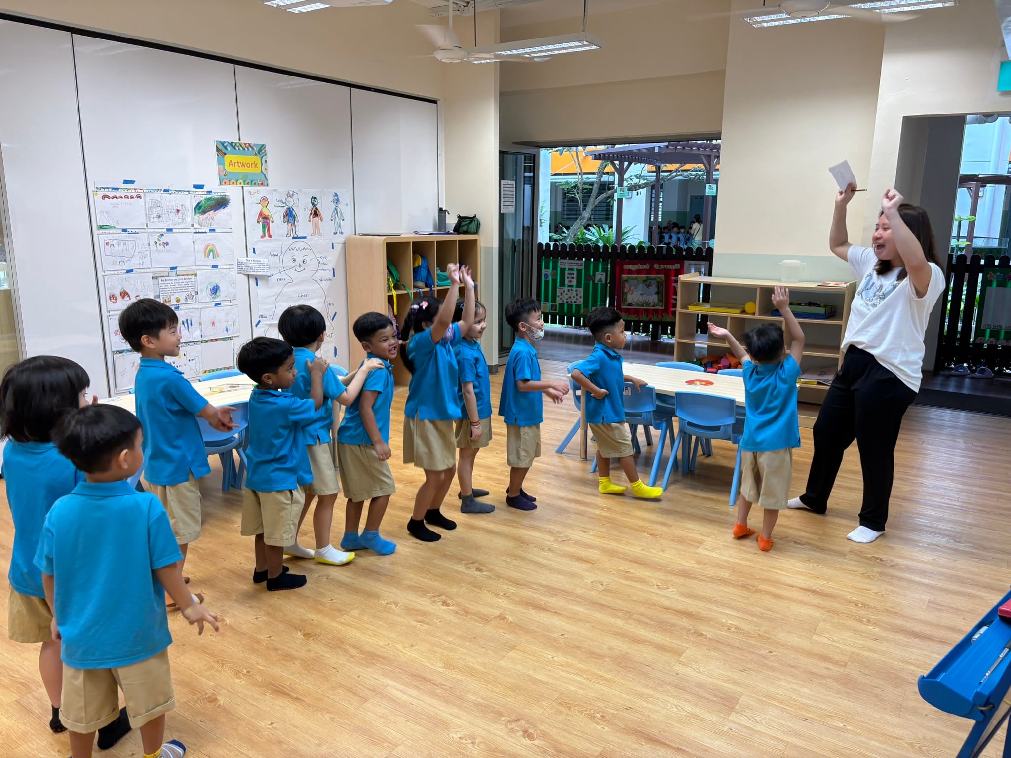 A teacher leads young children in blue uniforms in an energetic activity inside a bright classroom.