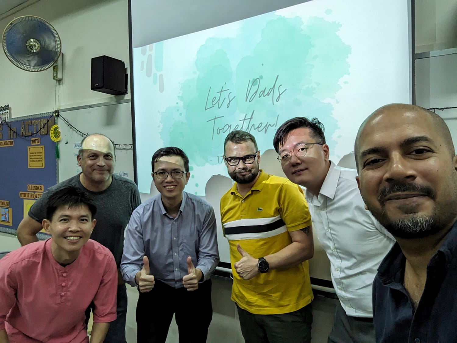 Six men smiling and giving thumbs up in front of a projection screen reading "Let's Dads Together" in a classroom.
