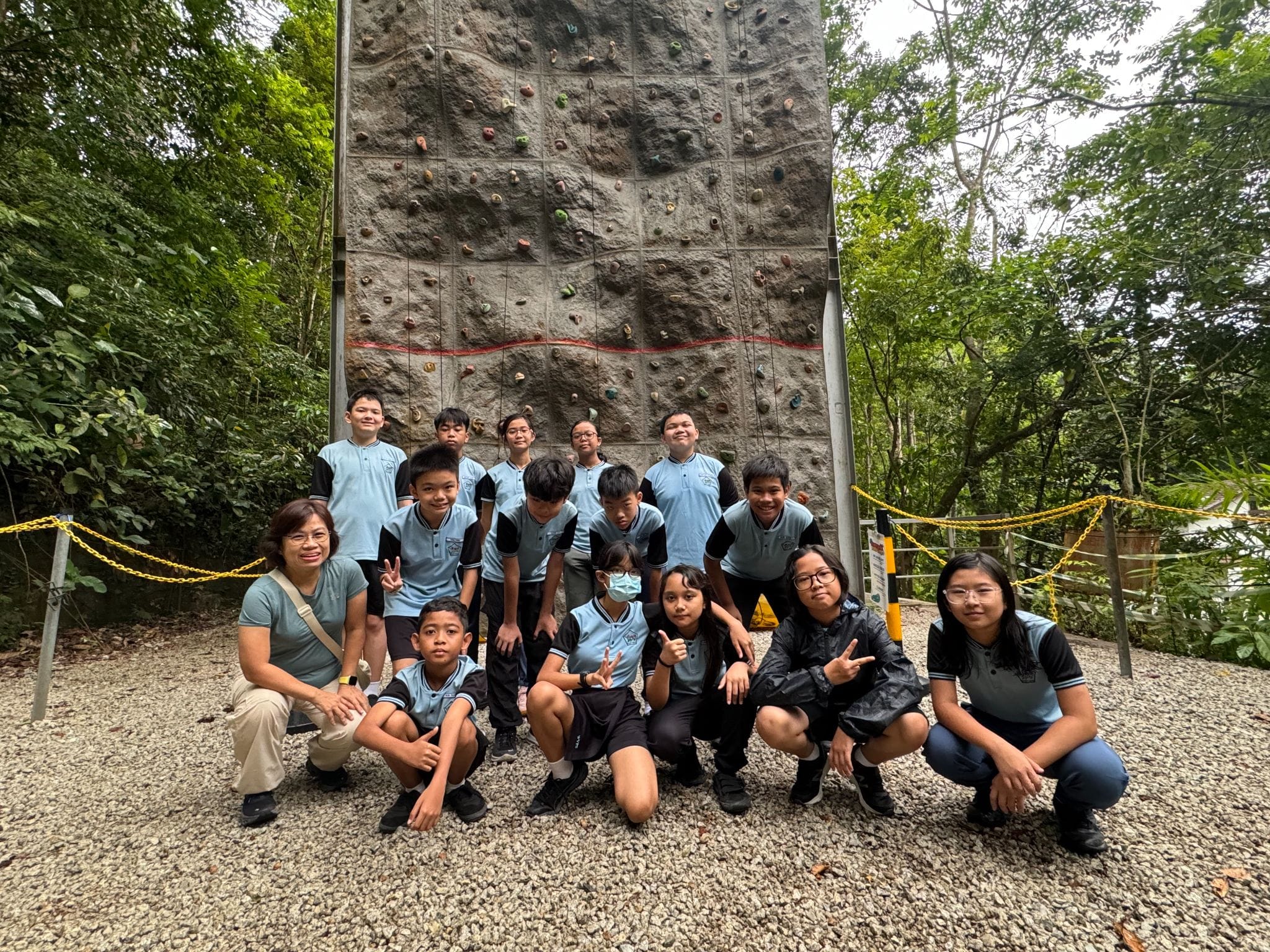 A group of students and teachers in light blue uniforms pose in front of a tall outdoor rock climbing wall surrounded by trees.
