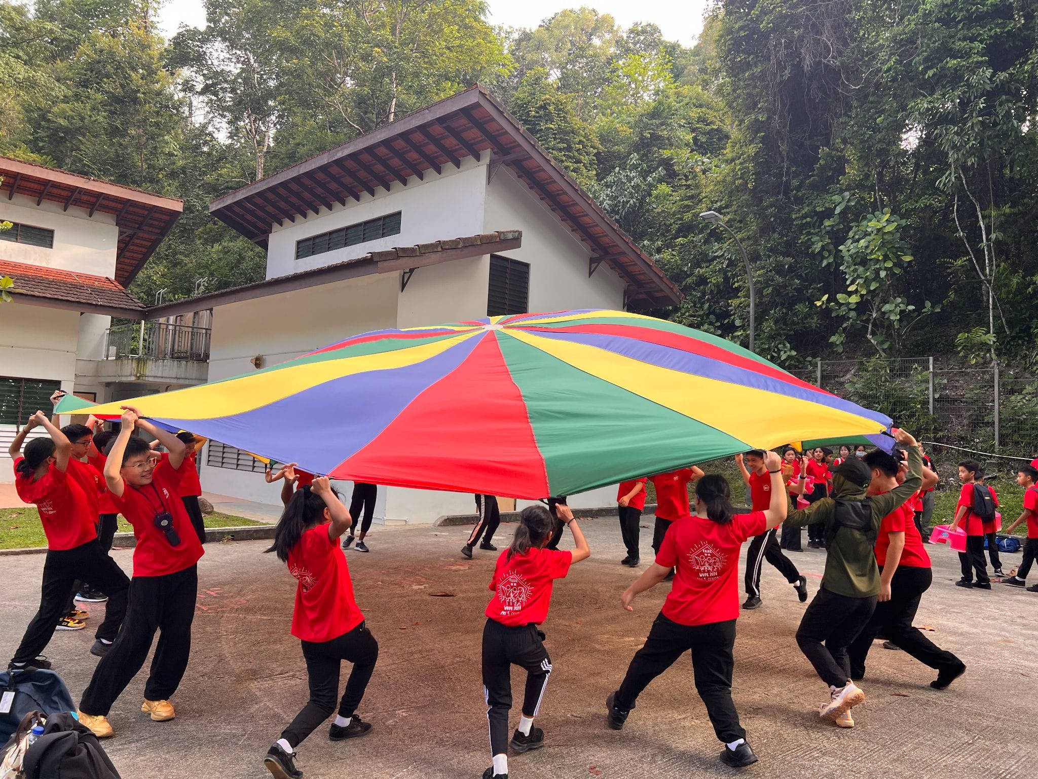 Children in red shirts play with a large colorful parachute outdoors near a white building surrounded by trees.
