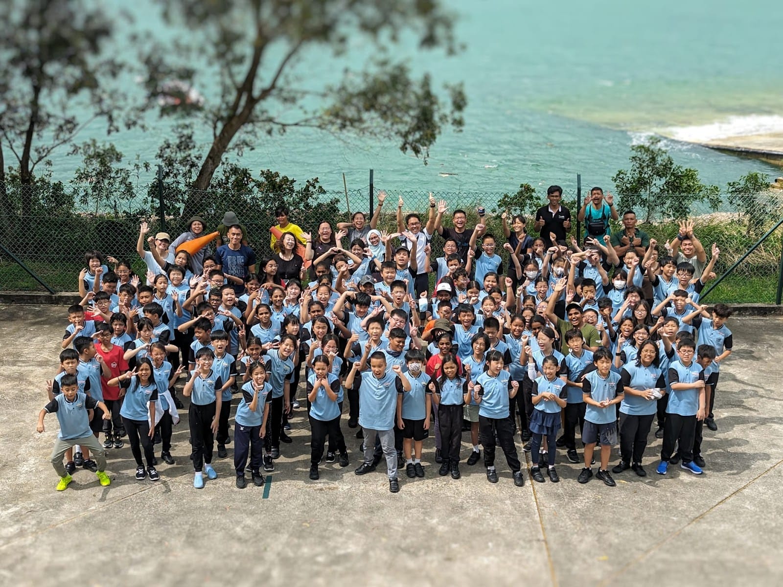 Large group of children in light blue uniforms posing cheerfully outdoors near a turquoise sea.