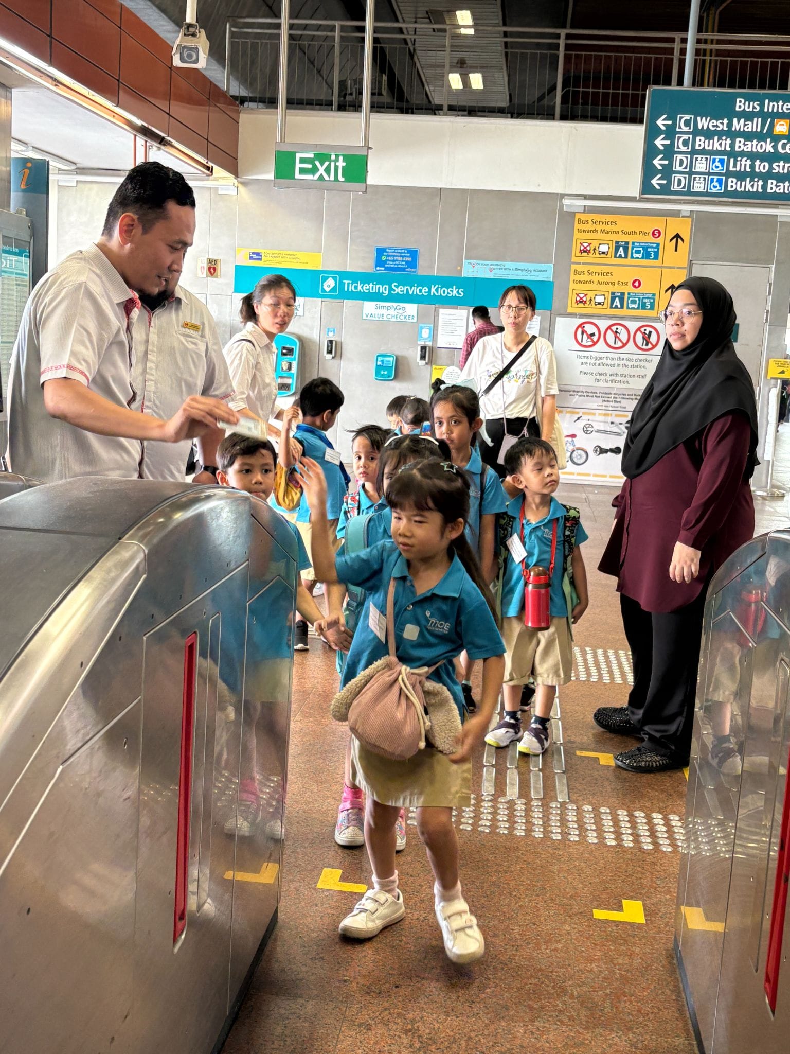 Young children in blue uniforms pass through fare gates at a Singapore MRT station, guided by adults nearby.