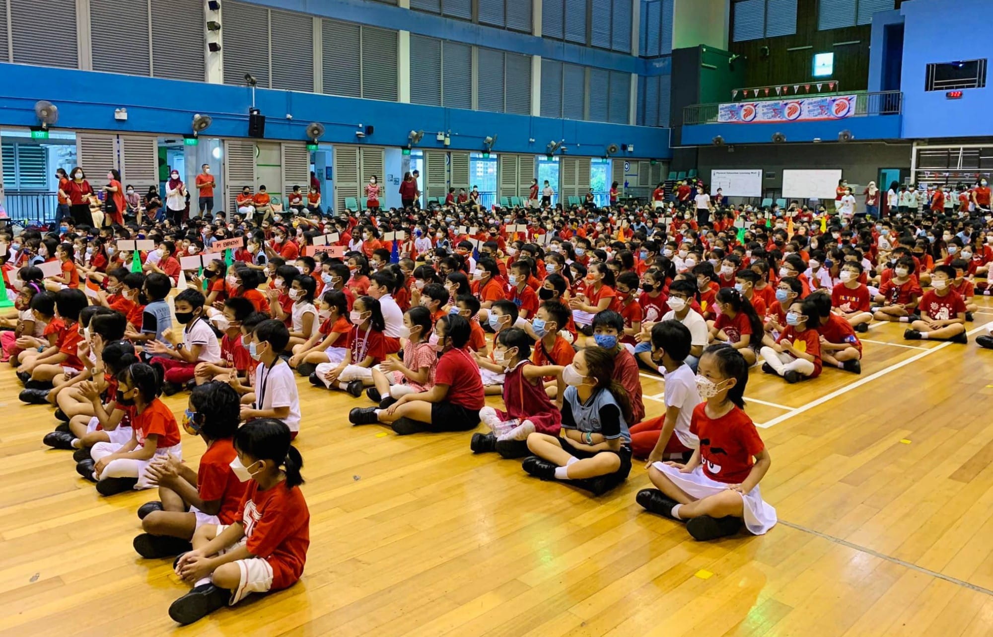 Hundreds of masked children in red and white uniforms sit on a gymnasium floor during a school assembly.