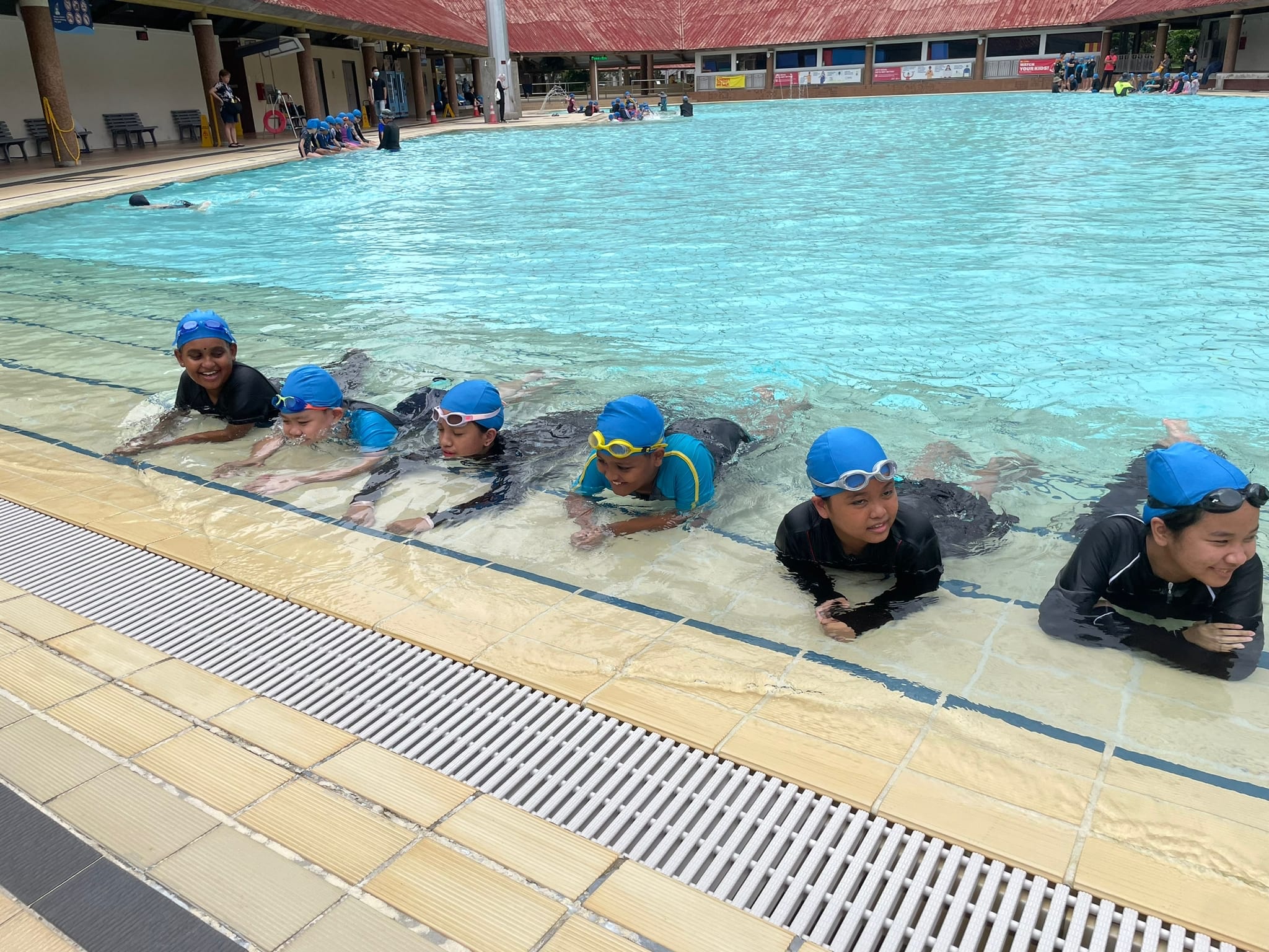 Children in blue swim caps and goggles line up at the edge of an outdoor pool during a swimming lesson.