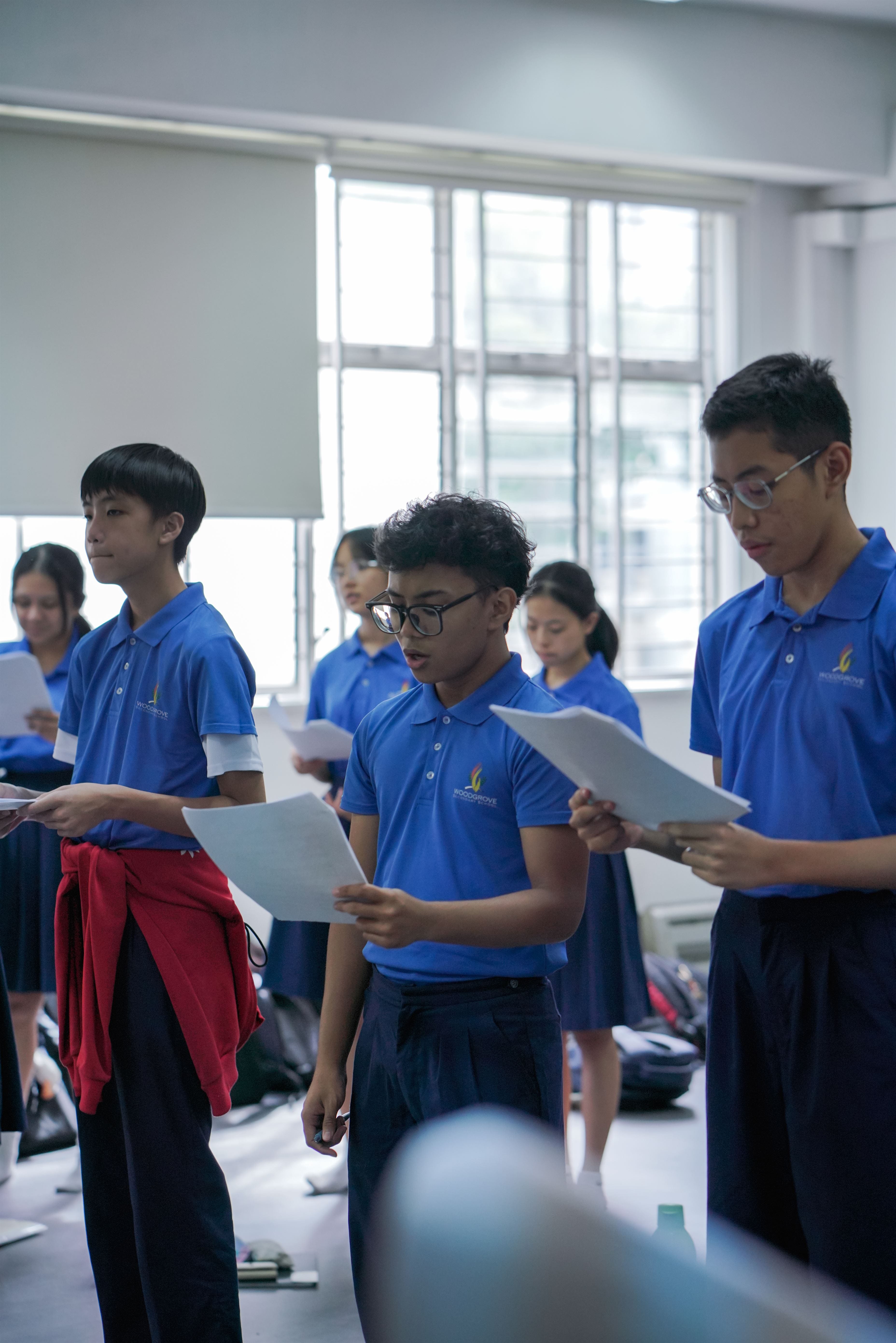 Several students in blue school uniforms stand in a classroom holding and reading from white paper sheets.