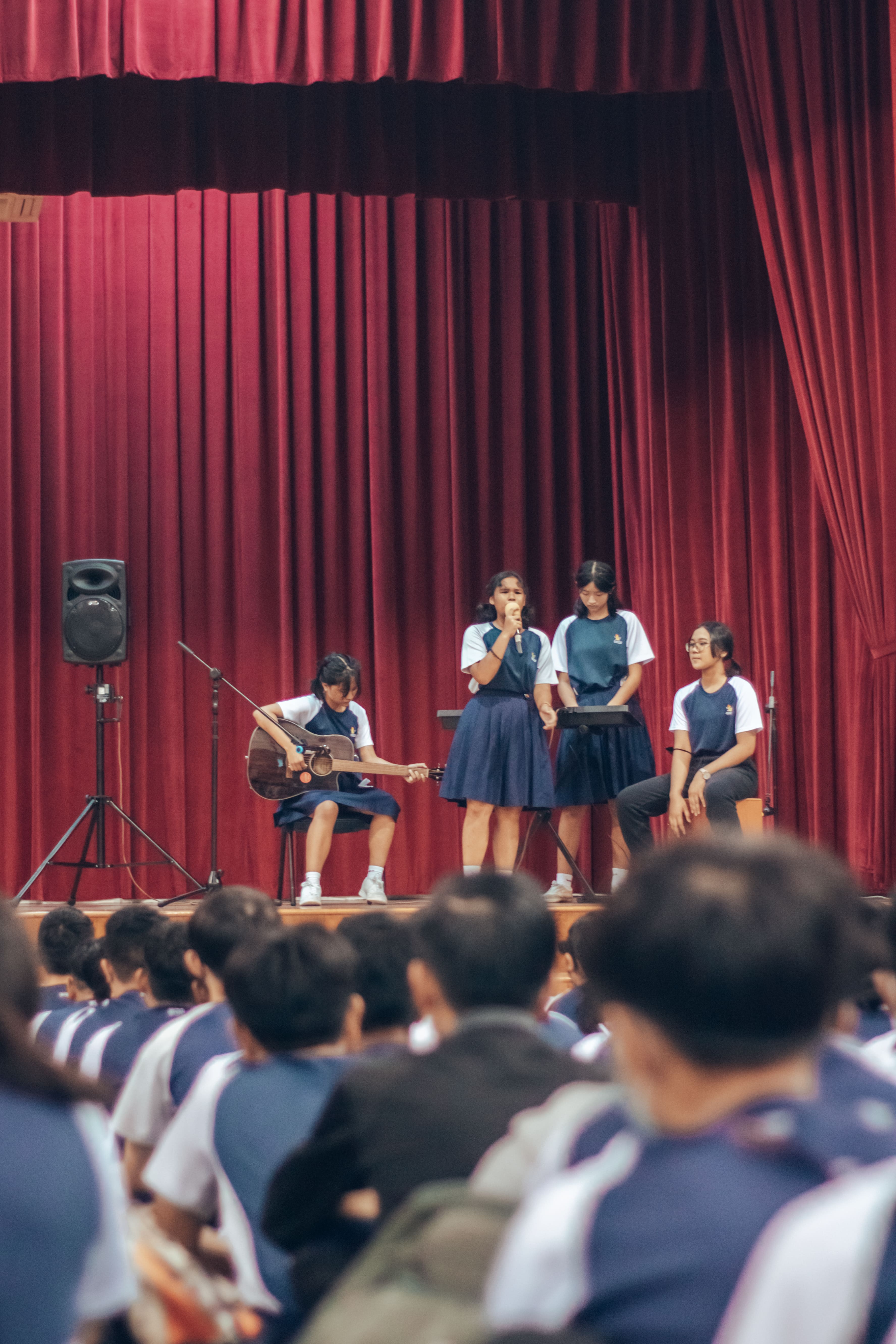 Four students in school uniforms perform on a stage with red curtains, one playing guitar and one singing into a microphone.