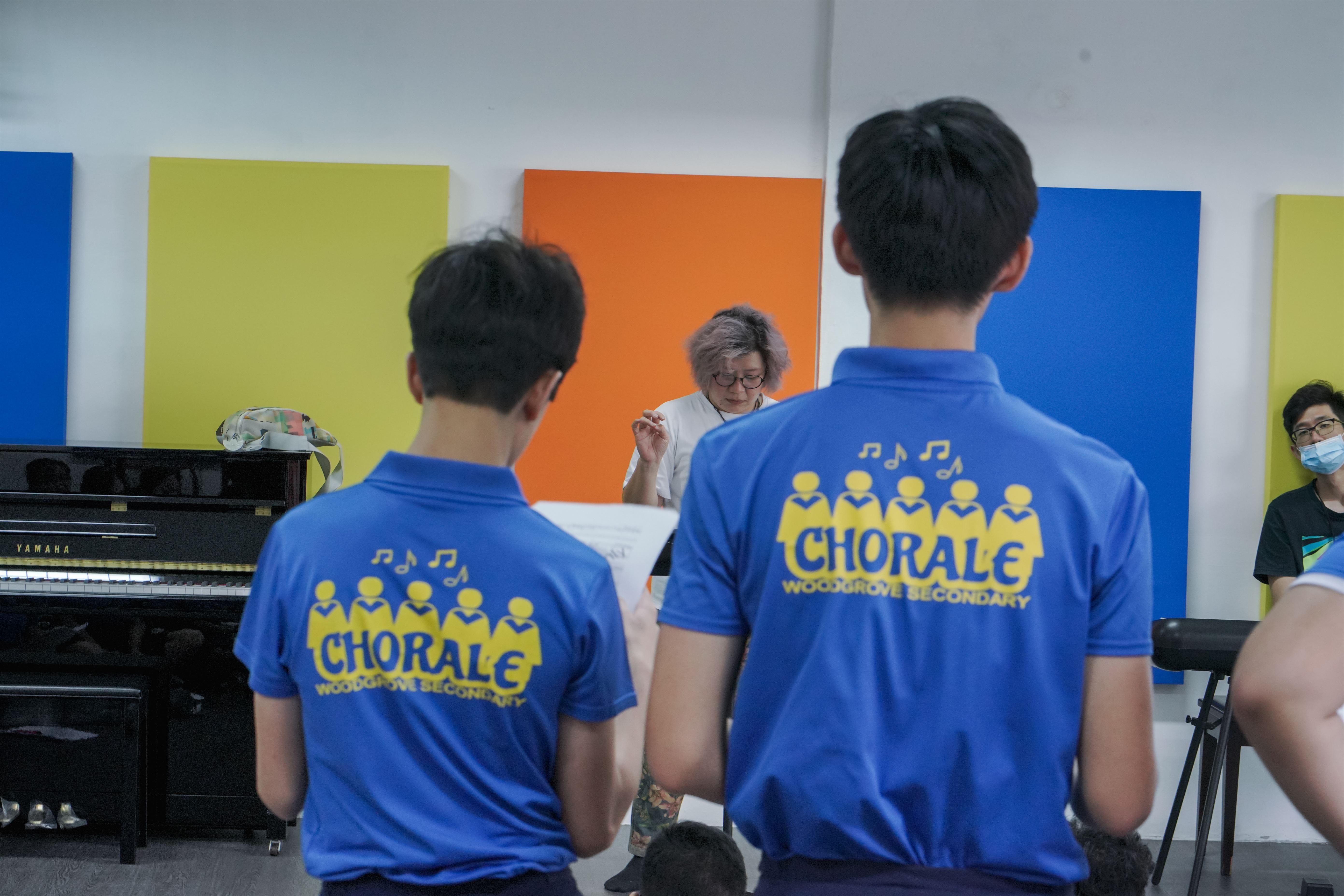 Two students in blue Woodgrove Secondary Chorale shirts face a conductor in a rehearsal room with a Yamaha piano.