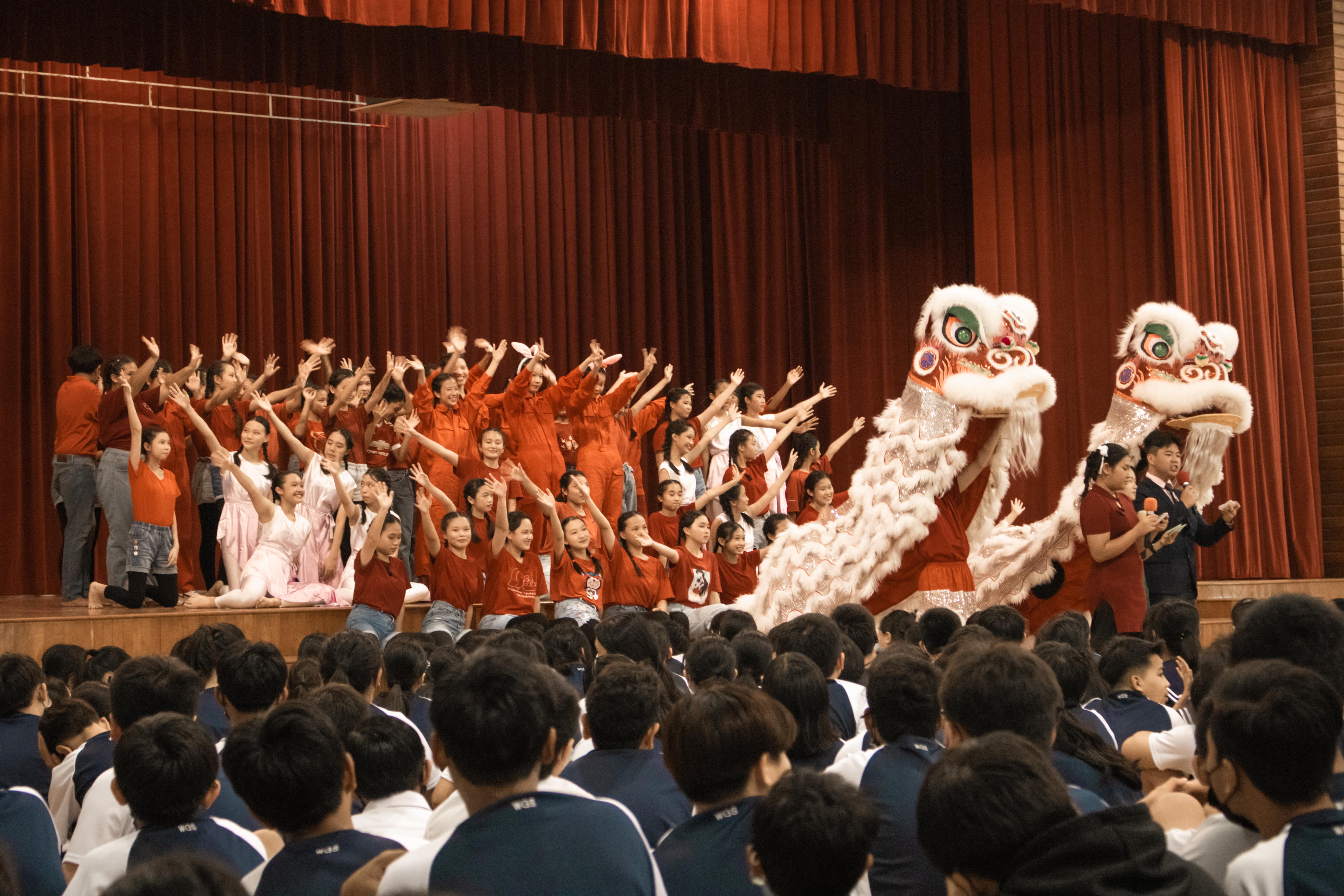 Students in red perform a Chinese lion dance on a stage with red curtains before a large seated audience.