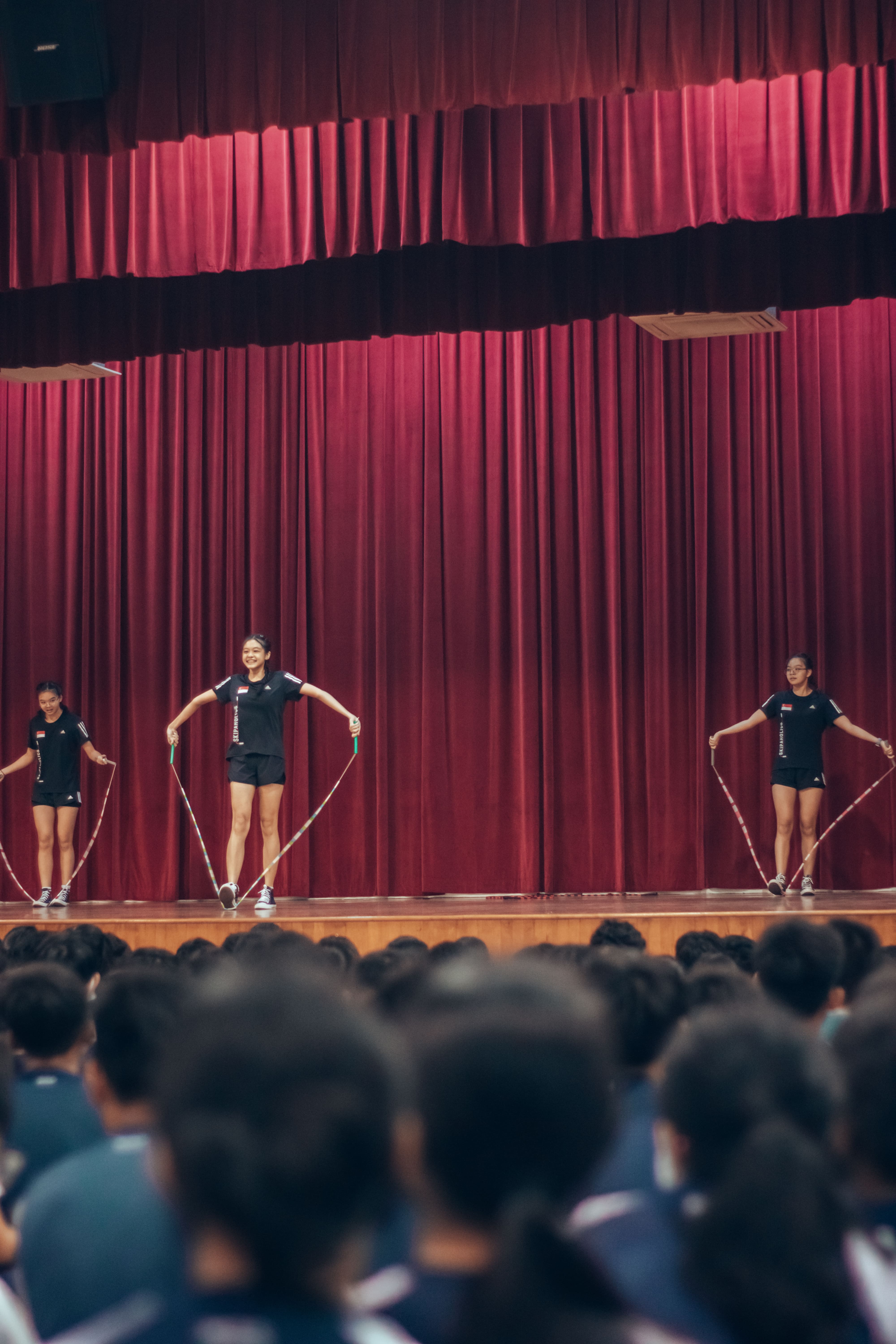Three girls in black uniforms perform jump rope on a stage with red curtains before a seated audience.