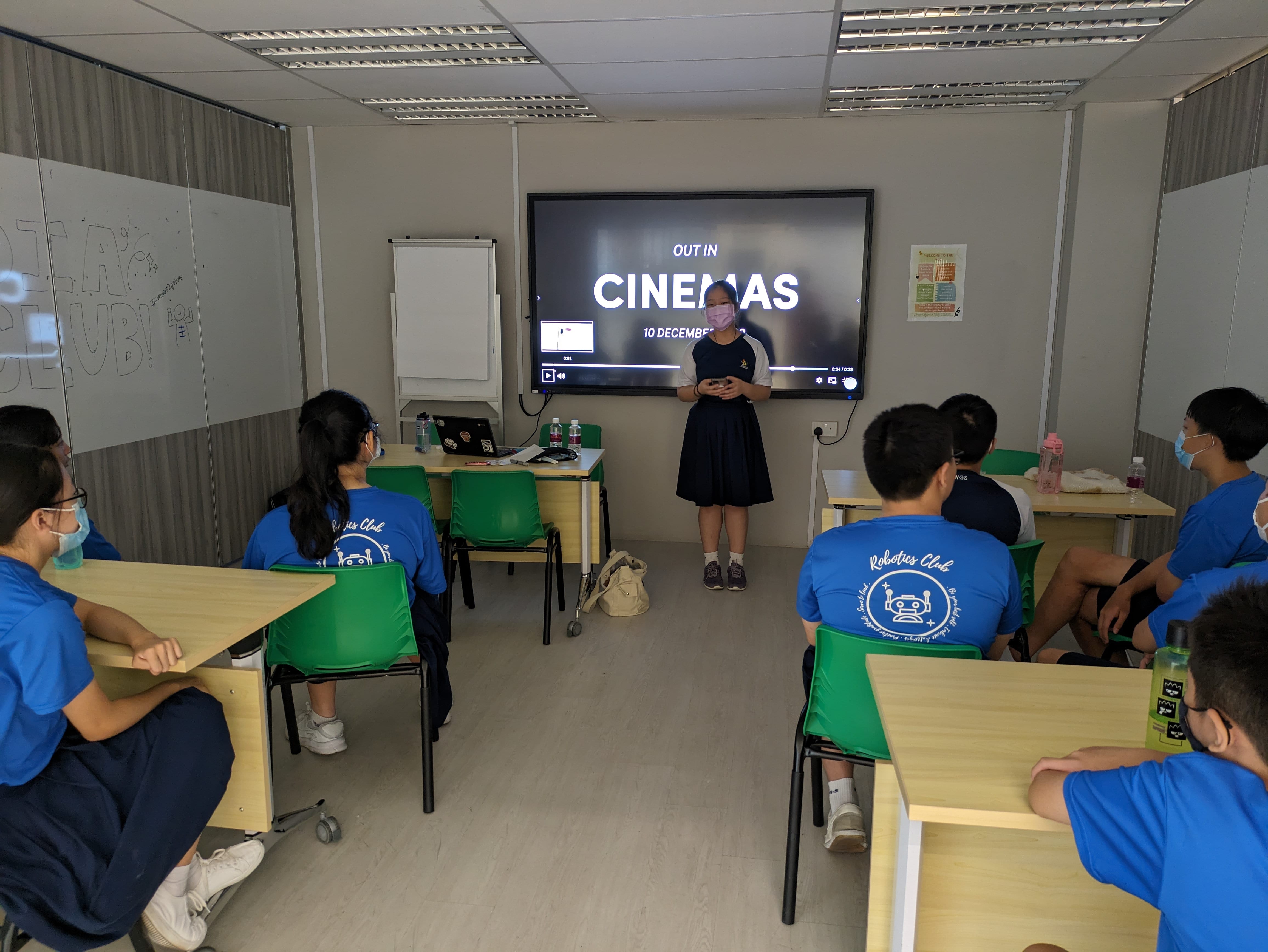 A student presents to masked classmates in blue Robotics Club shirts, with a screen showing "Out in Cinemas 10 December."