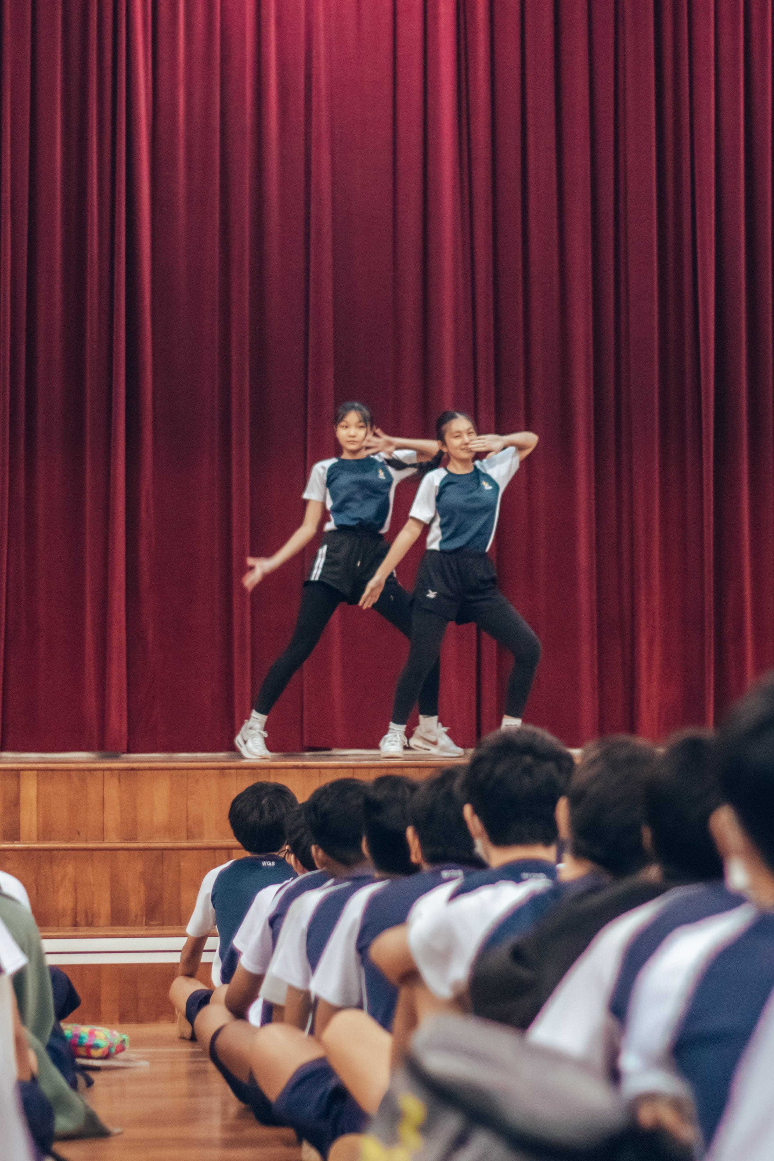 Two girls in matching uniforms dance on a wooden stage before a red curtain while students sit watching on the floor.