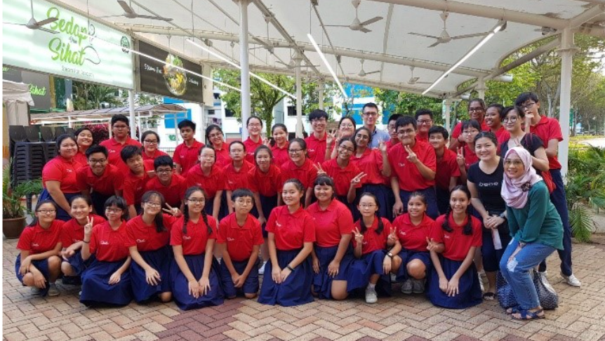 Large group of students in red shirts and navy skirts posing outdoors under a canopy near a food stall.