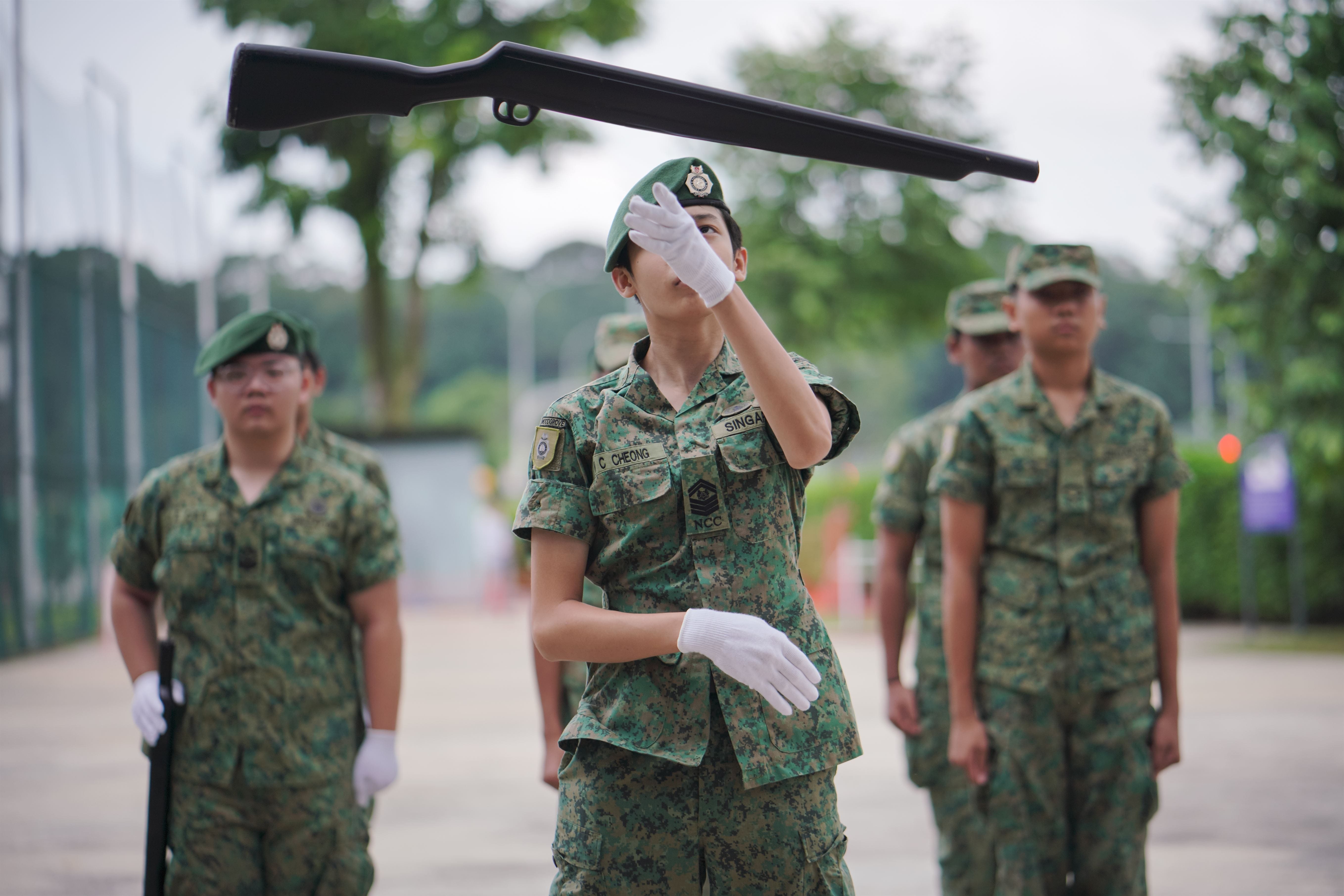 A uniformed cadet in green beret balances a rifle overhead during a drill, with other cadets standing behind.