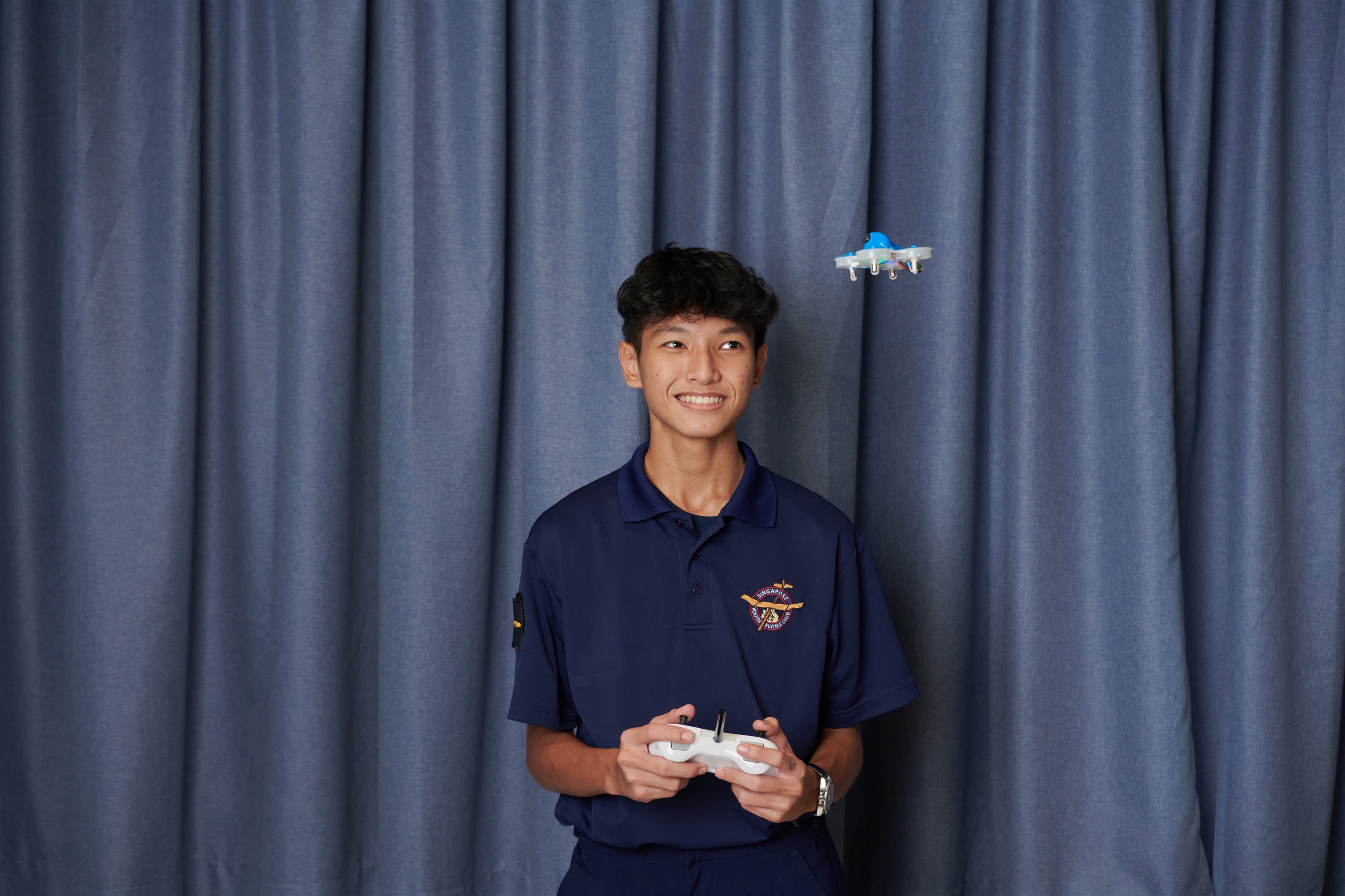 Student in Youth Flying Club uniform holds a drone remote controller while a small drone hovers beside him.