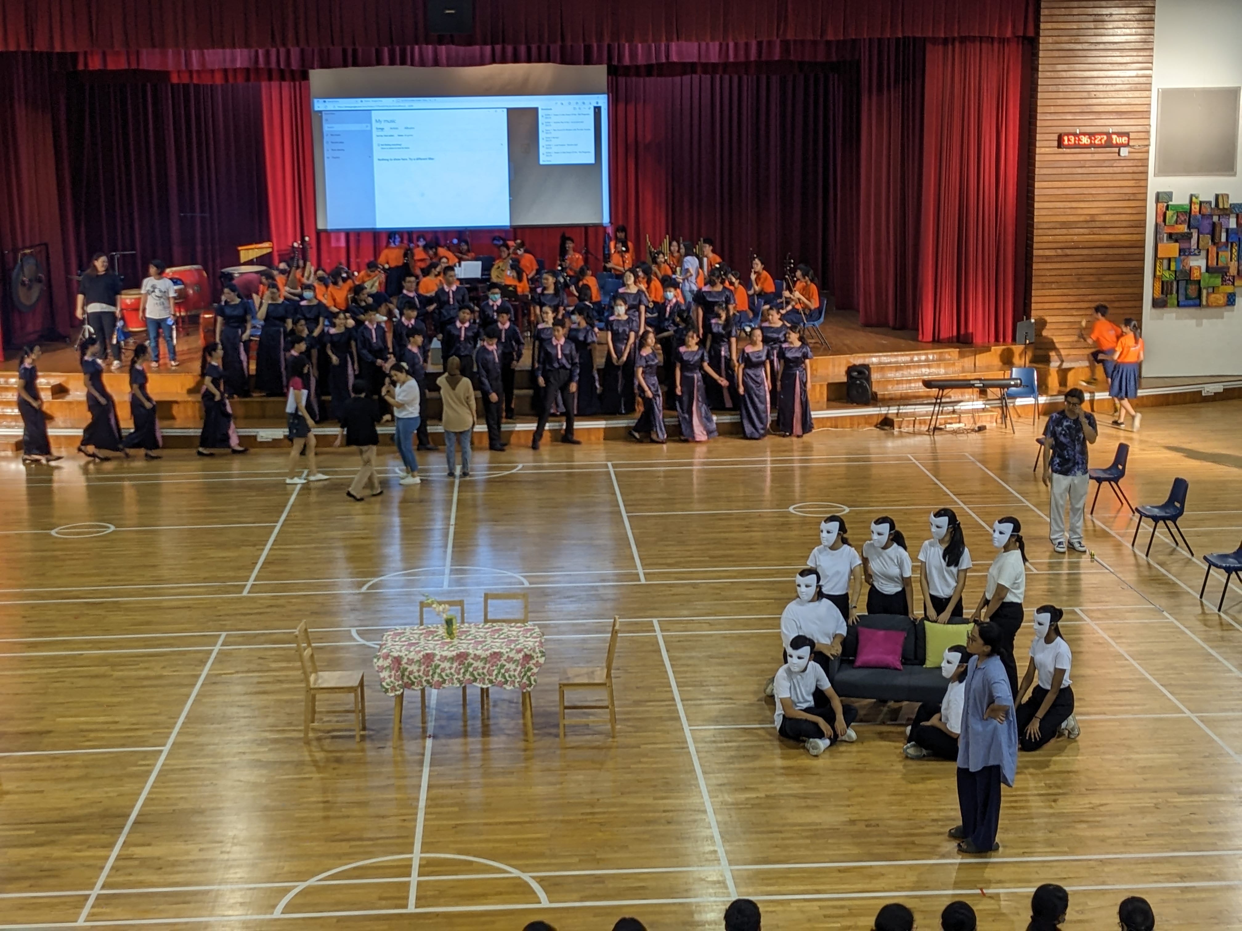 Students perform in a school hall, with a choir on stage and masked performers on the gymnasium floor near a small table.