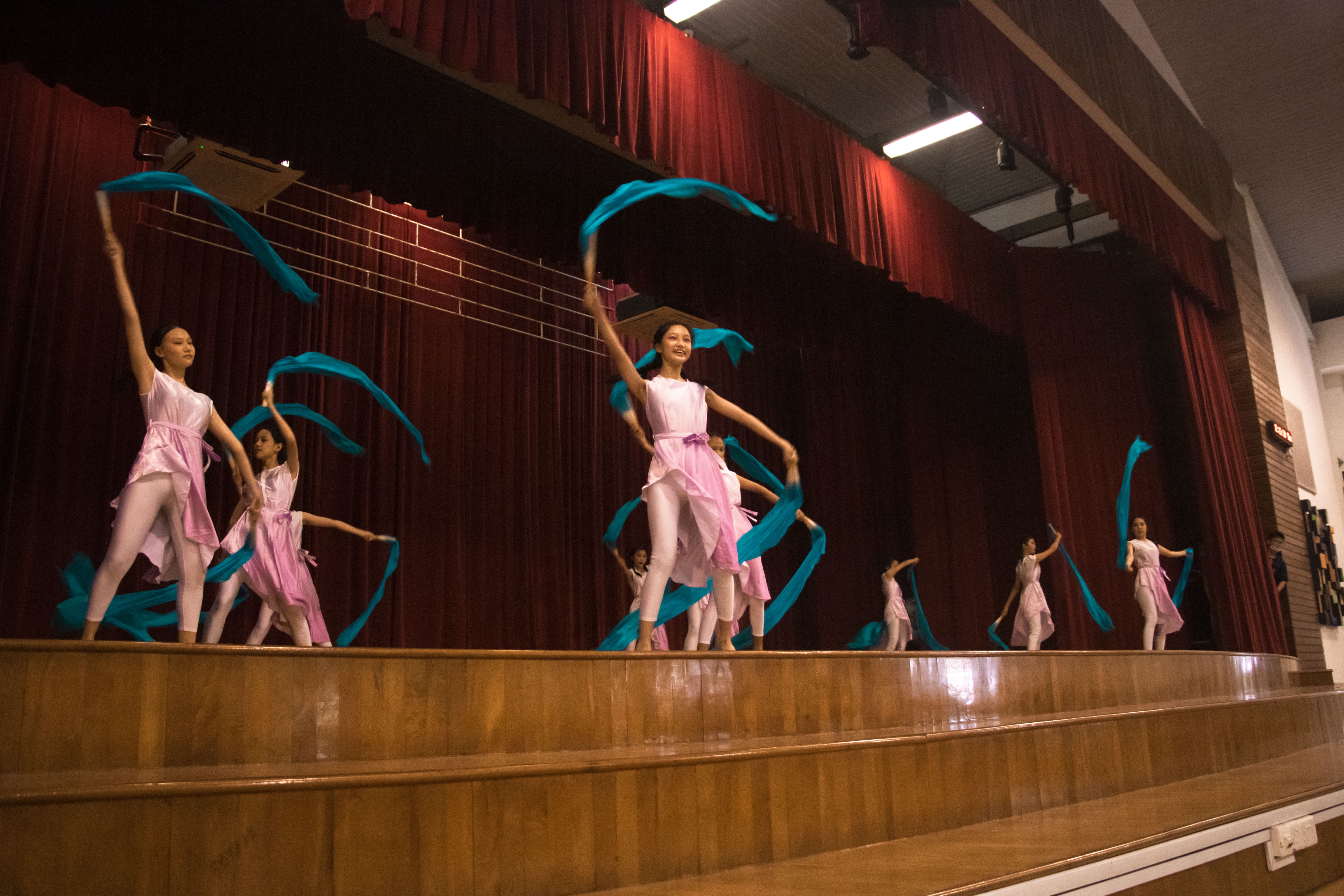 Several young female dancers in pink costumes wave teal ribbons on a wooden stage with red curtains behind them.
