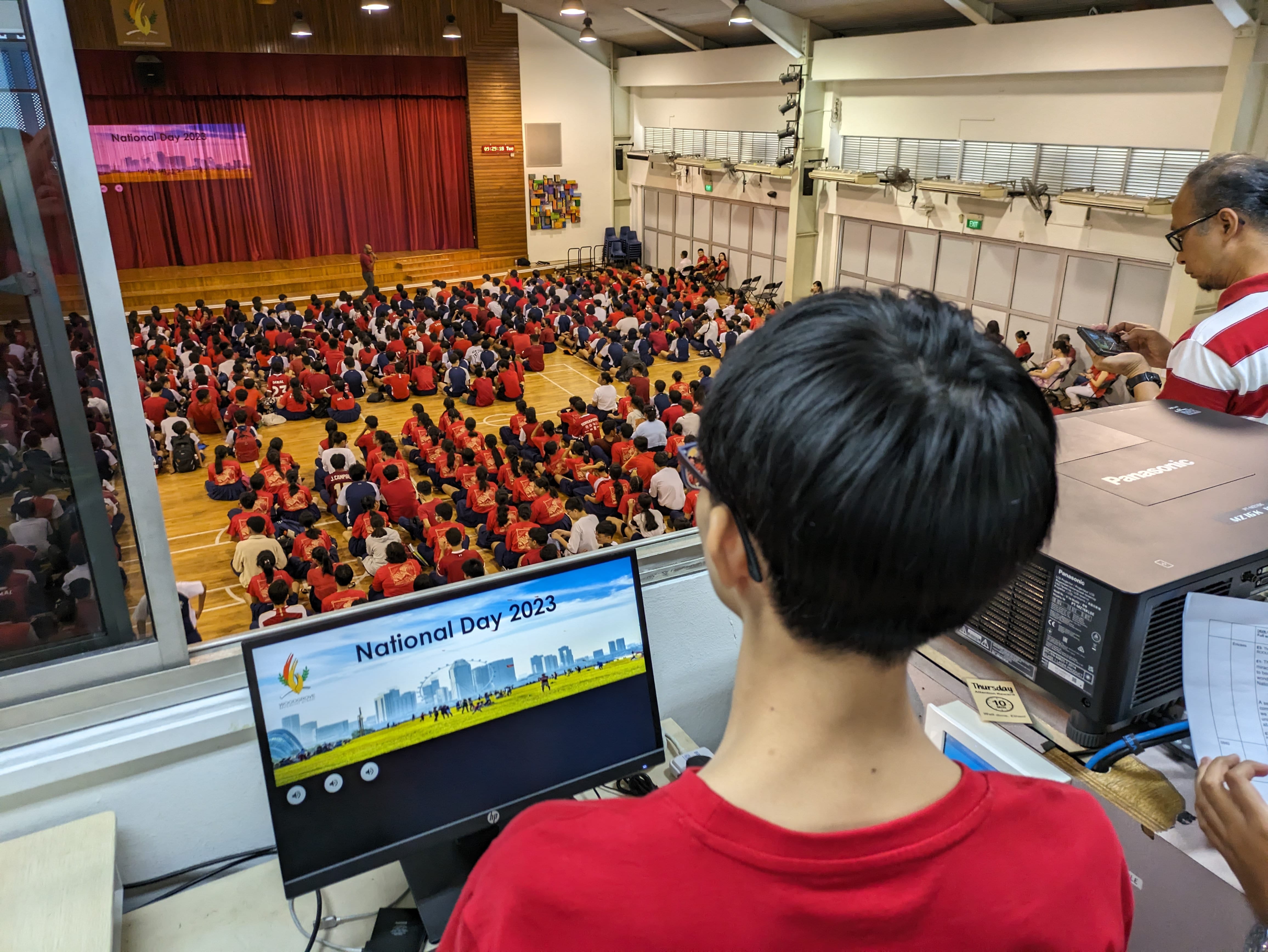 A person in red oversees a school National Day 2023 assembly, with hundreds of students seated on a hall floor.