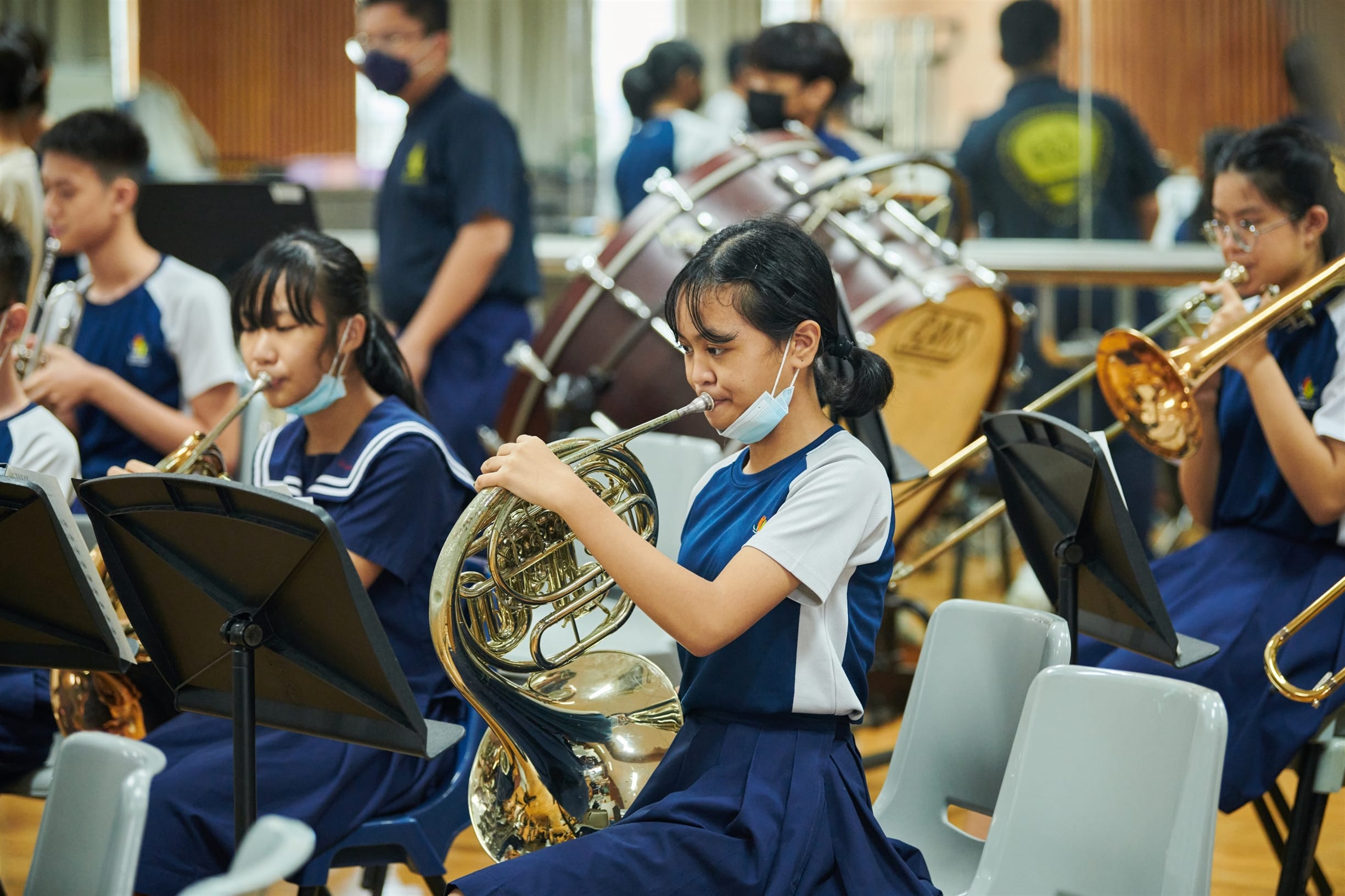 School students in uniforms playing brass instruments including a French horn and trombone during band rehearsal.