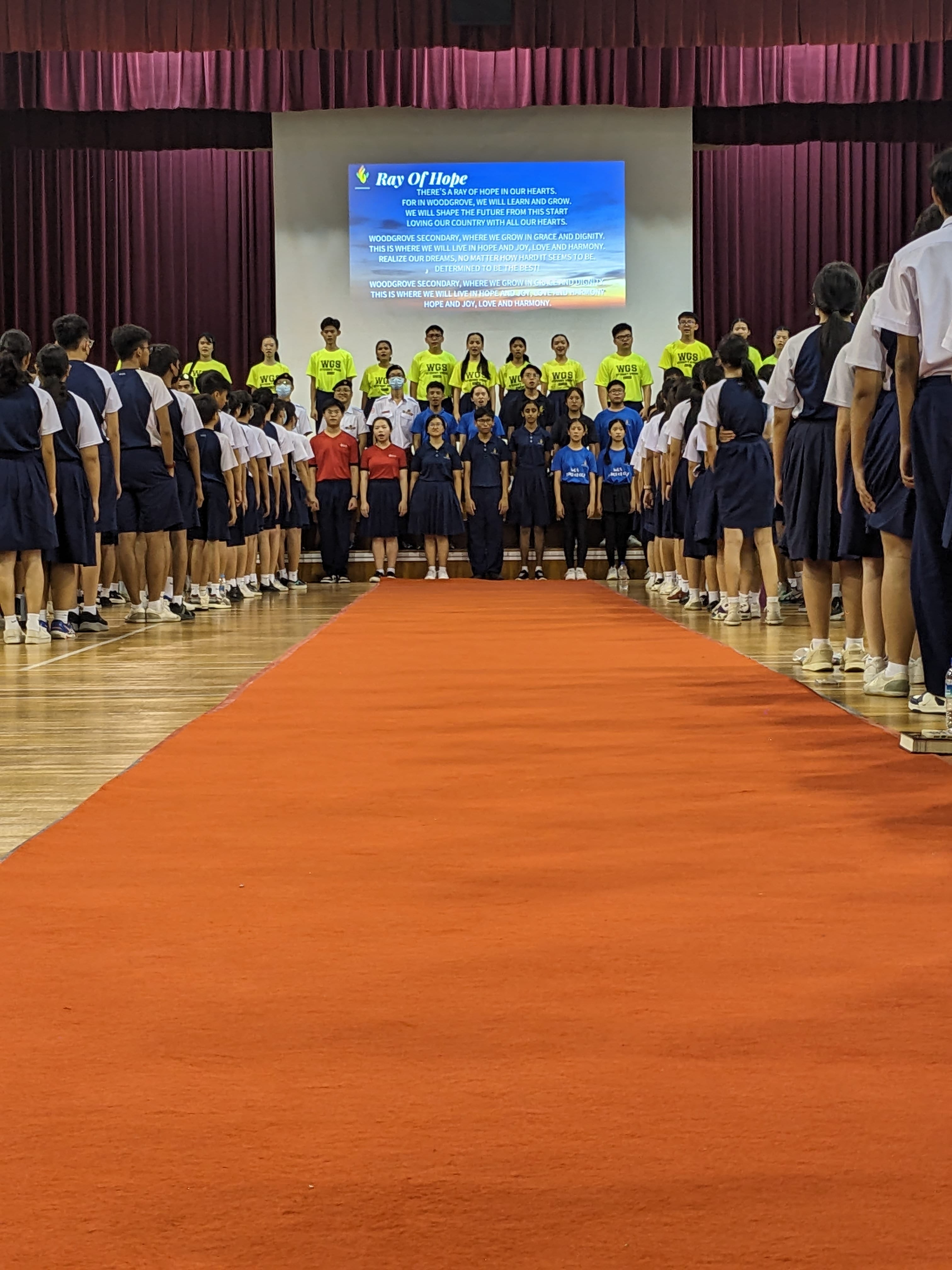 Students in uniforms stand in a school hall along a red carpet, facing a screen displaying "Ray Of Hope" lyrics.