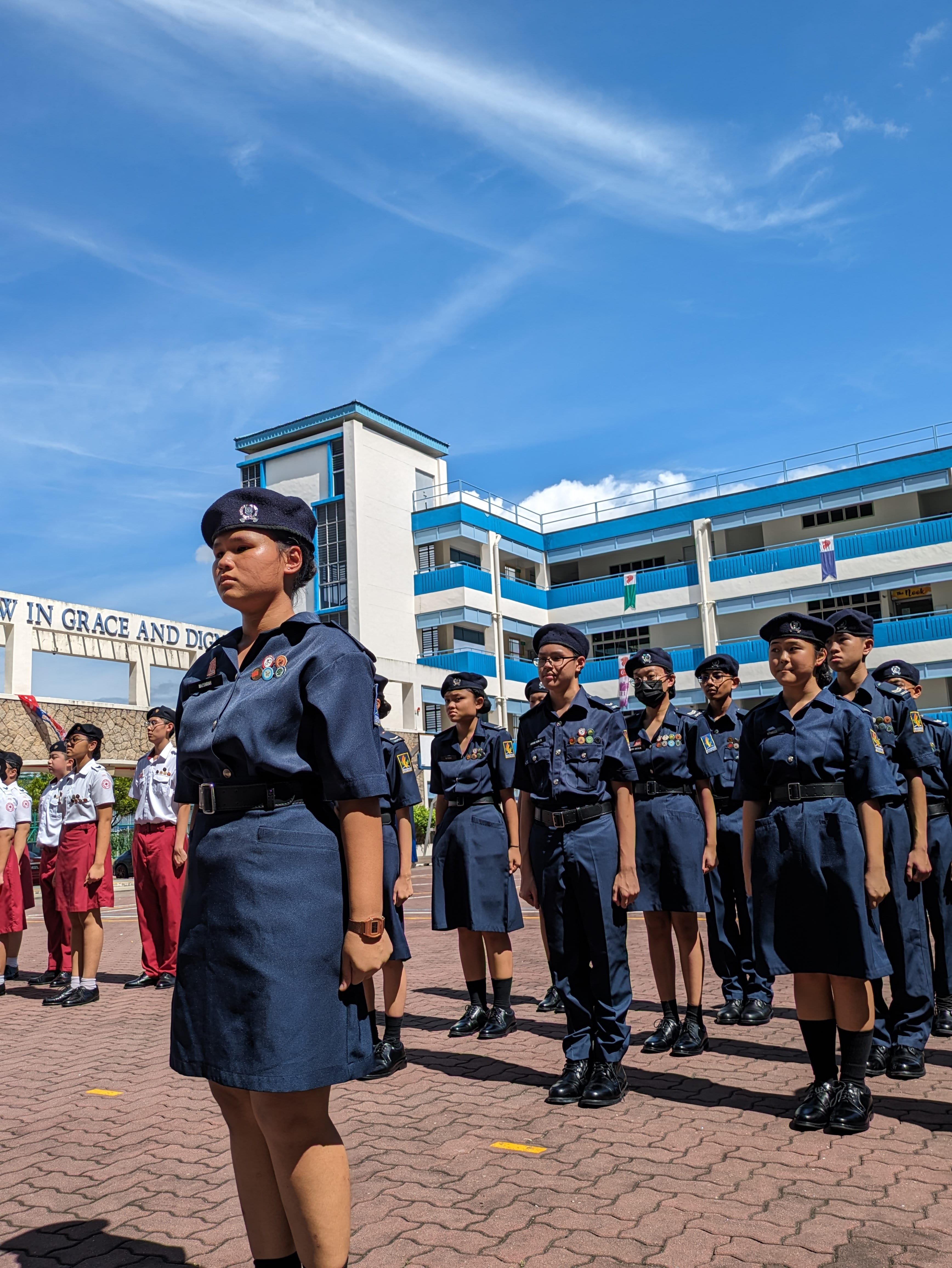 Students in navy blue uniforms and berets stand in formation on a school courtyard under a blue sky.